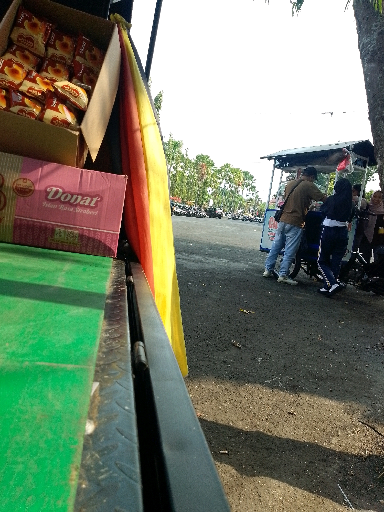 The image captures a scene outside, possibly in the city of Jember, Indonesia, with a focus on a food cart and its surroundings. In the foreground, a green surface is visible, possibly part of a vehicle or a stall, with a black metal edge. To the right of this green surface, a section of a cart or stall is seen, draped with a vibrantly colored fabric, featuring yellow, orange, and red hues.
Further back, a food cart stands open, with a roof supported by metal poles. A man wearing a brown shirt and light blue jeans is interacting with someone at the cart. He has a bag slung over his shoulder. Beside him, a woman in a black hijab and athletic pants is also at the cart. Another woman in a hijab, smiling, is visible behind them. A motorcycle is parked next to the cart.
In the background, a line of parked motorcycles suggests a public area, possibly a park or a marketplace. Numerous palm trees line the horizon, indicating a tropical or subtropical climate. The ground is paved with asphalt, and there are scattered leaves and debris. The sky is bright and appears to be daytime. The overall impression is that of a casual outdoor setting where food is being sold.