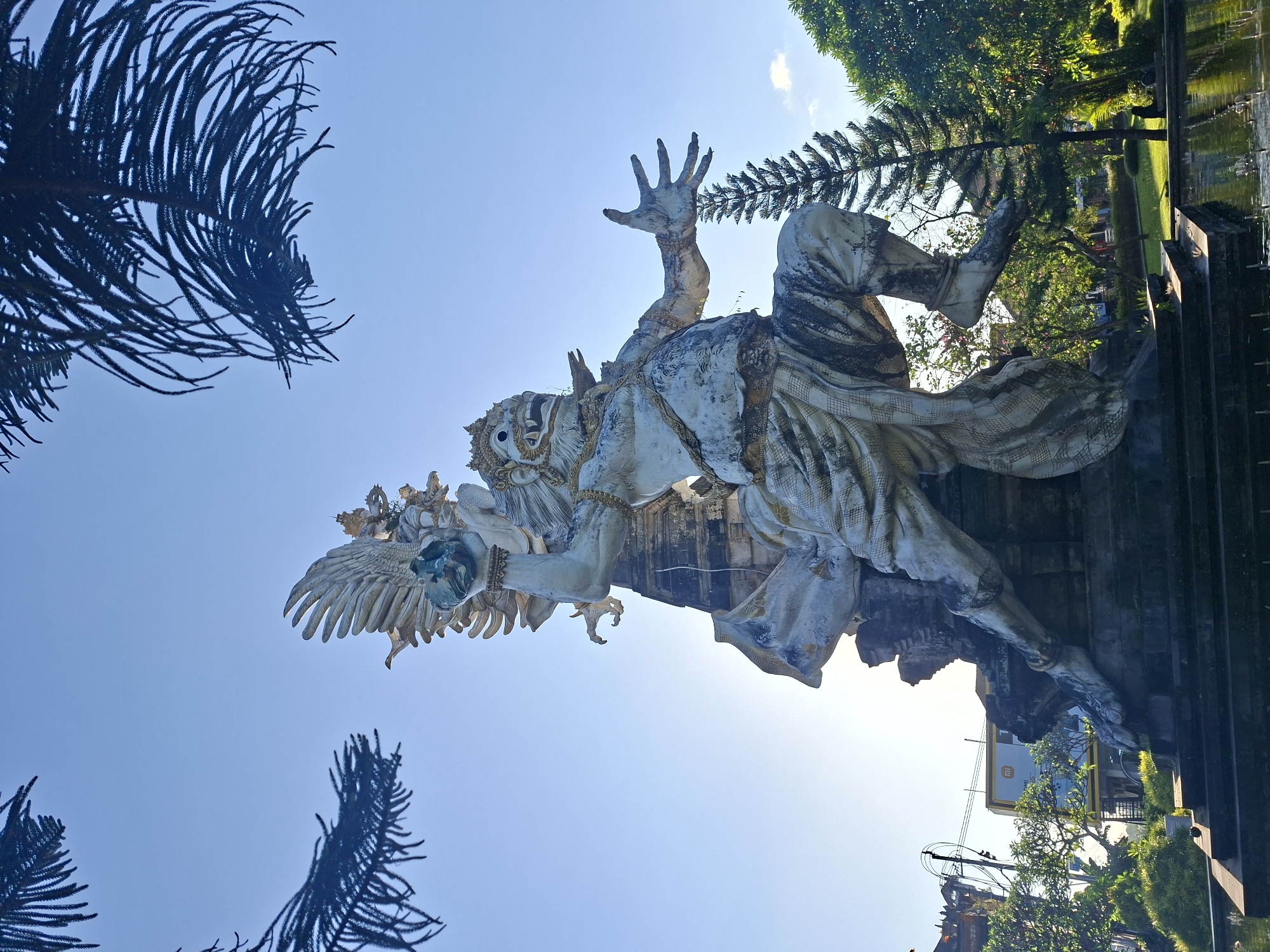 A large, weathered stone statue of a Garuda, a mythical bird-like creature from Hindu mythology, stands majestically against a clear blue sky. The statue depicts the Garuda in a dynamic pose, with one arm raised holding a globe, wings spread, and its other arm extended outwards. The creature has a fierce expression with a detailed face, adorned with a crown and jewelry. It is dressed in traditional Balinese attire, with flowing pants and a sash. The statue is situated in a park-like setting, with lush green trees and a body of water in the foreground, reflecting the sunlight. The statue is located at the Taman Festival Bali, an abandoned cultural park in Gianyar, Indonesia. The sun is bright, casting shadows and highlighting the textures of the stone.