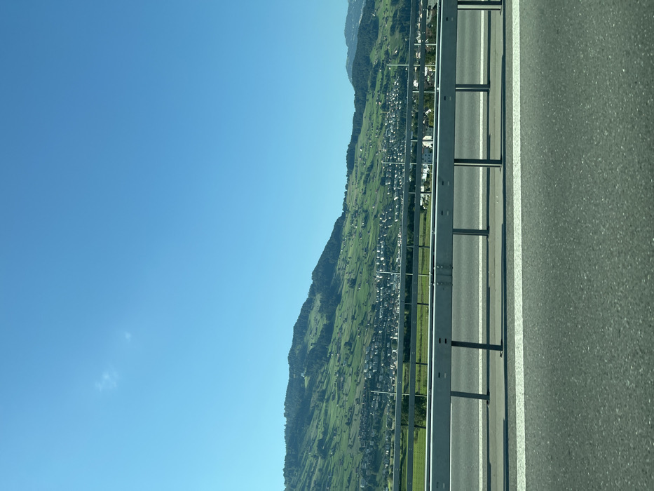 The image presents a daytime, exterior view of a landscape in Buochs, Switzerland, captured from a roadway. The foreground features a dark asphalt surface with two parallel white lines. A light grey, segmented metal guardrail extends horizontally across the lower-mid ground, supported by vertical posts that cast shadows. Beyond the guardrail, a flat, vibrant green field is visible, transitioning into a cluster of light-colored buildings with dark roofs, representing a town situated at the base of a large, green-covered hill. This hill, sparsely dotted with trees and fields, occupies the central and left background. The upper two-fifths of the frame consist of a clear, bright blue sky, with a faint, wispy white cloud in the upper-left quadrant. No individuals are visible.