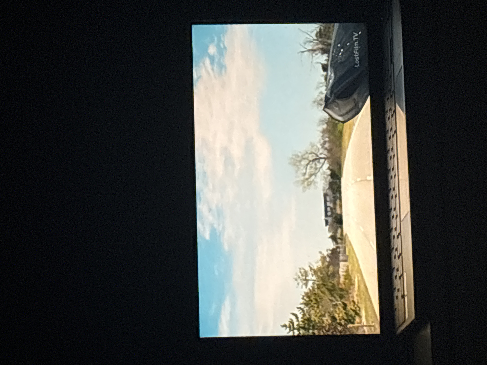 The image shows a screen displaying a scene with a road curving to the right, flanked by vegetation. The sky is visible with a mix of blue and white clouds. A dark object partially obstructs the view at the top of the road. The screen itself has the text "LostFilm.TV" in the upper right corner. The surrounding environment is dark, suggesting the image is taken of a screen in a darkened room. There is no information to say where in the city of Budva, Montenegro that the content on the screen is of, nor is there anything to say it is of Budva, Montenegro.
