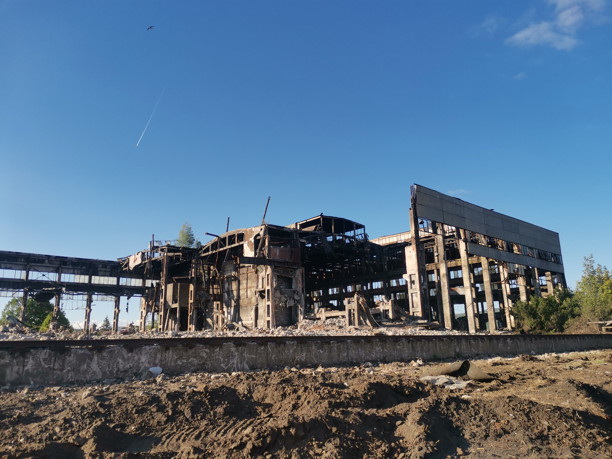 The image depicts the ruins of a large industrial building against a clear blue sky. The building is severely damaged, with a charred and collapsed central section and partially standing skeletal remains of its framework. Debris and rubble are scattered around the base of the structure, covering a dirt and gravel-strewn foreground. In the sky, a contrail and a bird are visible.
While Zhivkovo, Bulgaria is mentioned, there is no information in the image to verify the location.