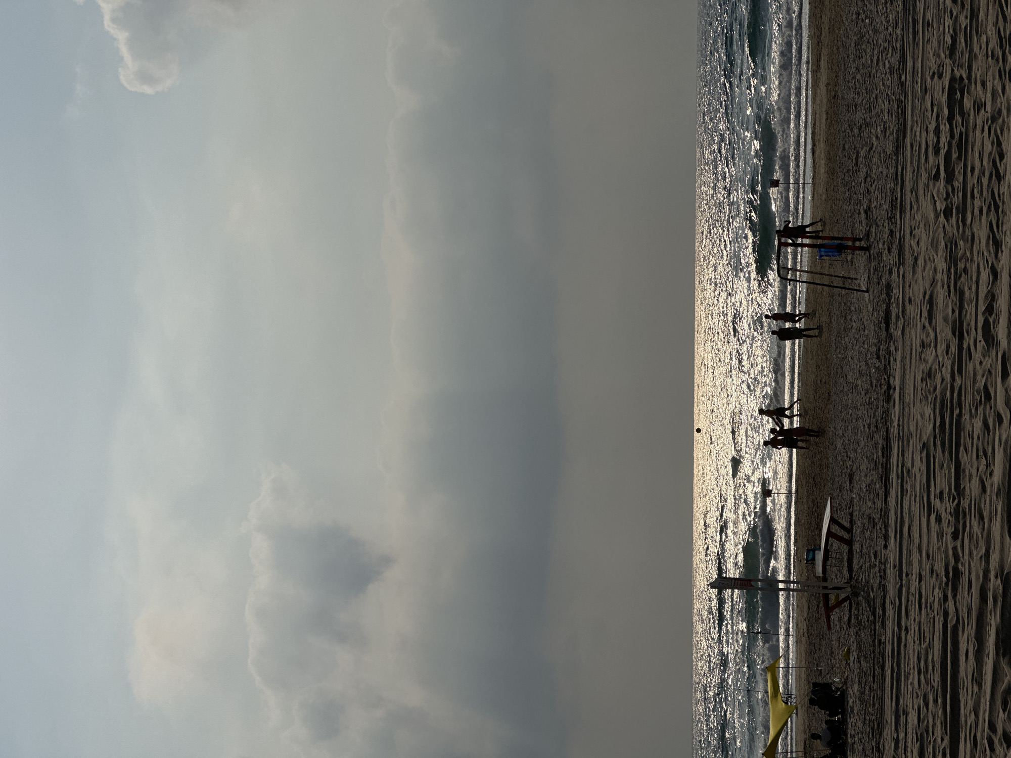 The image depicts a beach scene in Bat Yam, Israel, with the sun reflecting off the rippling ocean. In the foreground, the sandy beach is visible, with tire tracks imprinted in the sand. To the left, two yellow umbrellas provide shade over a group of seated individuals. A picnic table with benches stands in the middle ground.
Further down the beach, a group of silhouettes are engaged in a game, possibly soccer or volleyball, with a ball visible in the air. There is also a red and blue swing set structure. In the background, the ocean waves are breaking, and the horizon is obscured by a line of darker clouds. The overall lighting suggests it is either early morning or late afternoon.