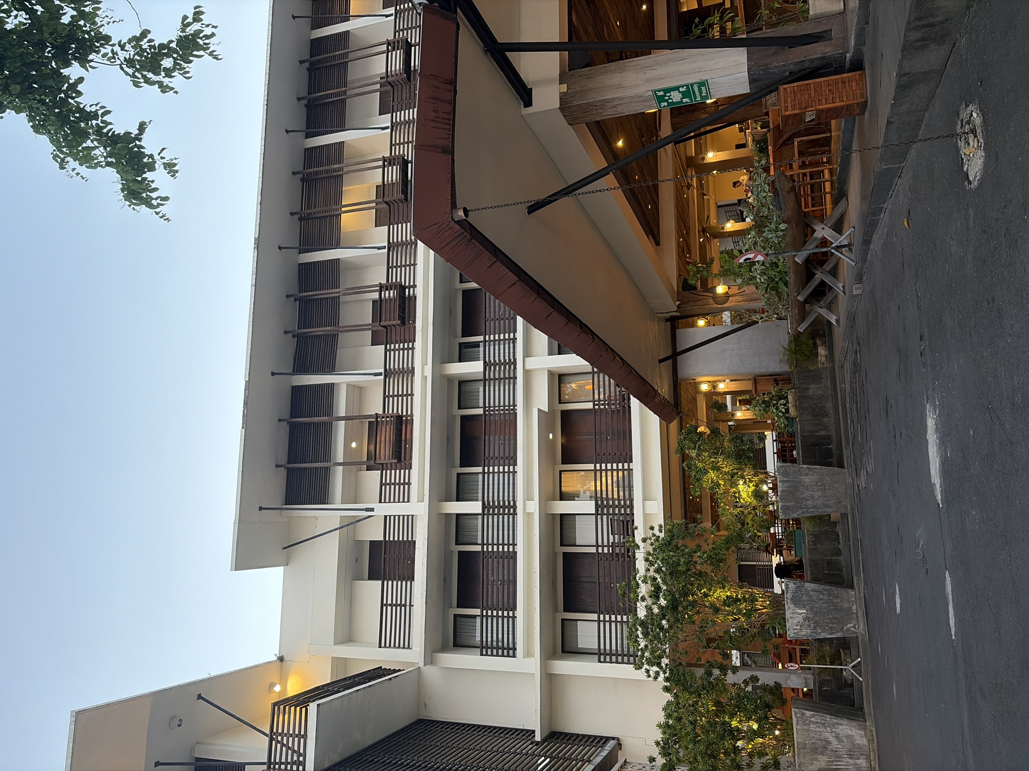An exterior shot captures a modern hotel building in Kuta, Indonesia, under a clear sky. The building features white walls, multiple balconies with dark railings, and wooden louvers above some of the windows. Large, concrete planters with lush green trees are situated in front of the building. A covered walkway extends from the building, leading to an open-air dining or lounge area with tables and chairs. A green sign indicating an "Assembly Point" is visible near the entrance. The ground in front of the hotel is paved with asphalt, showing some signs of wear. The lighting suggests it is either early morning or late afternoon.