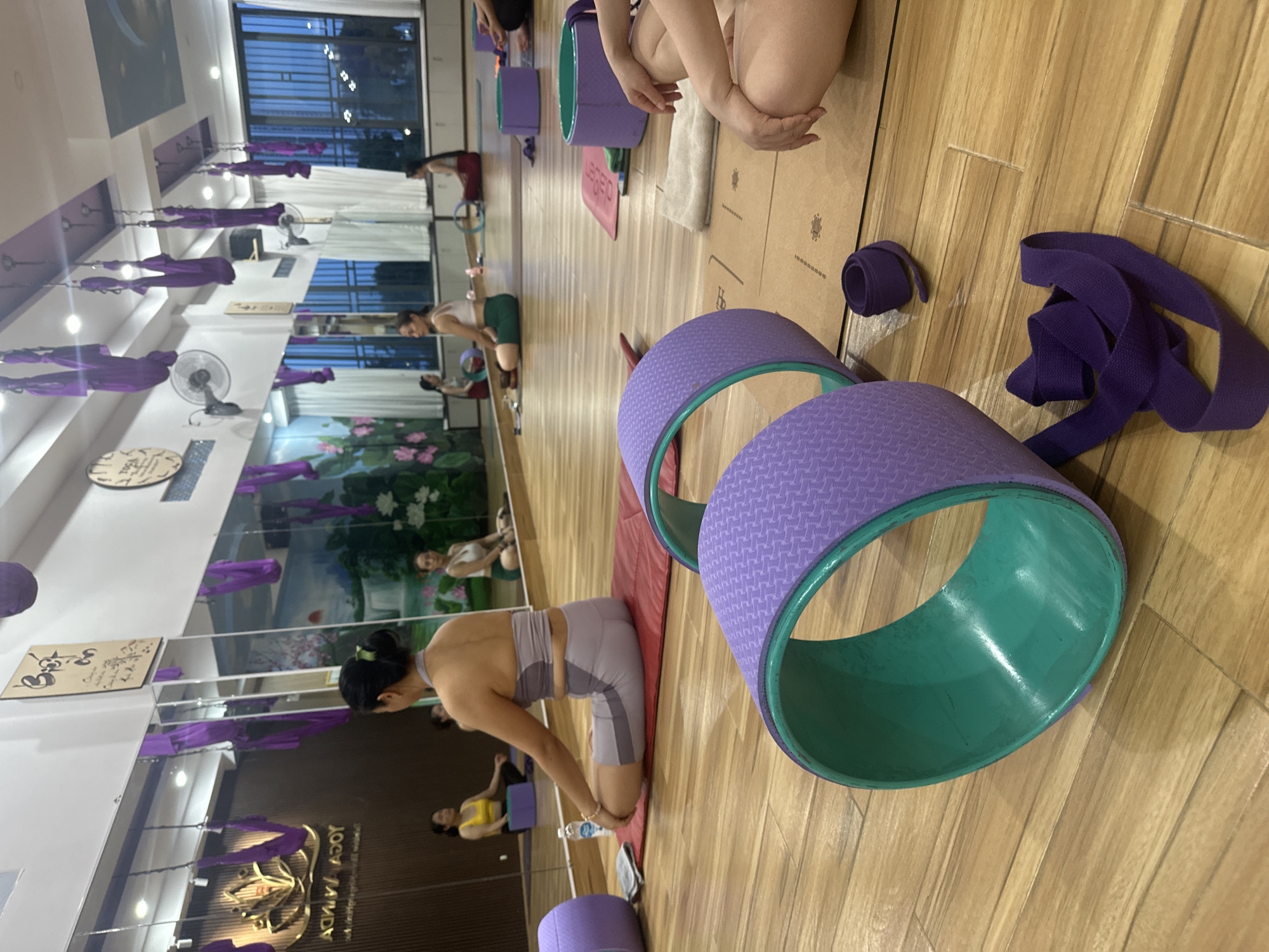 The image depicts the interior of a yoga studio in Bac Nha Trang Ward, Vietnam. The room is spacious with wooden floors and features large mirrors along one wall, reflecting the studio's amenities. Hanging from the ceiling are numerous purple aerial yoga hammocks, characteristic of this type of practice. Several women are present in the studio, engaged in or preparing for yoga. In the foreground, two large purple yoga wheels are visible, one with a teal inner rim, lying on the floor. A purple yoga strap is also present. In the mid-ground, a woman in a grey yoga outfit is seated on a red yoga mat in a meditative pose, with her legs crossed. Other women are seen in the reflection of the mirrors, also in seated yoga positions on mats. The studio is equipped with various yoga props, including yoga blocks and more yoga wheels, suggesting a functional space for yoga classes. The overall ambiance suggests a calm and focused environment for physical and mental well-being.