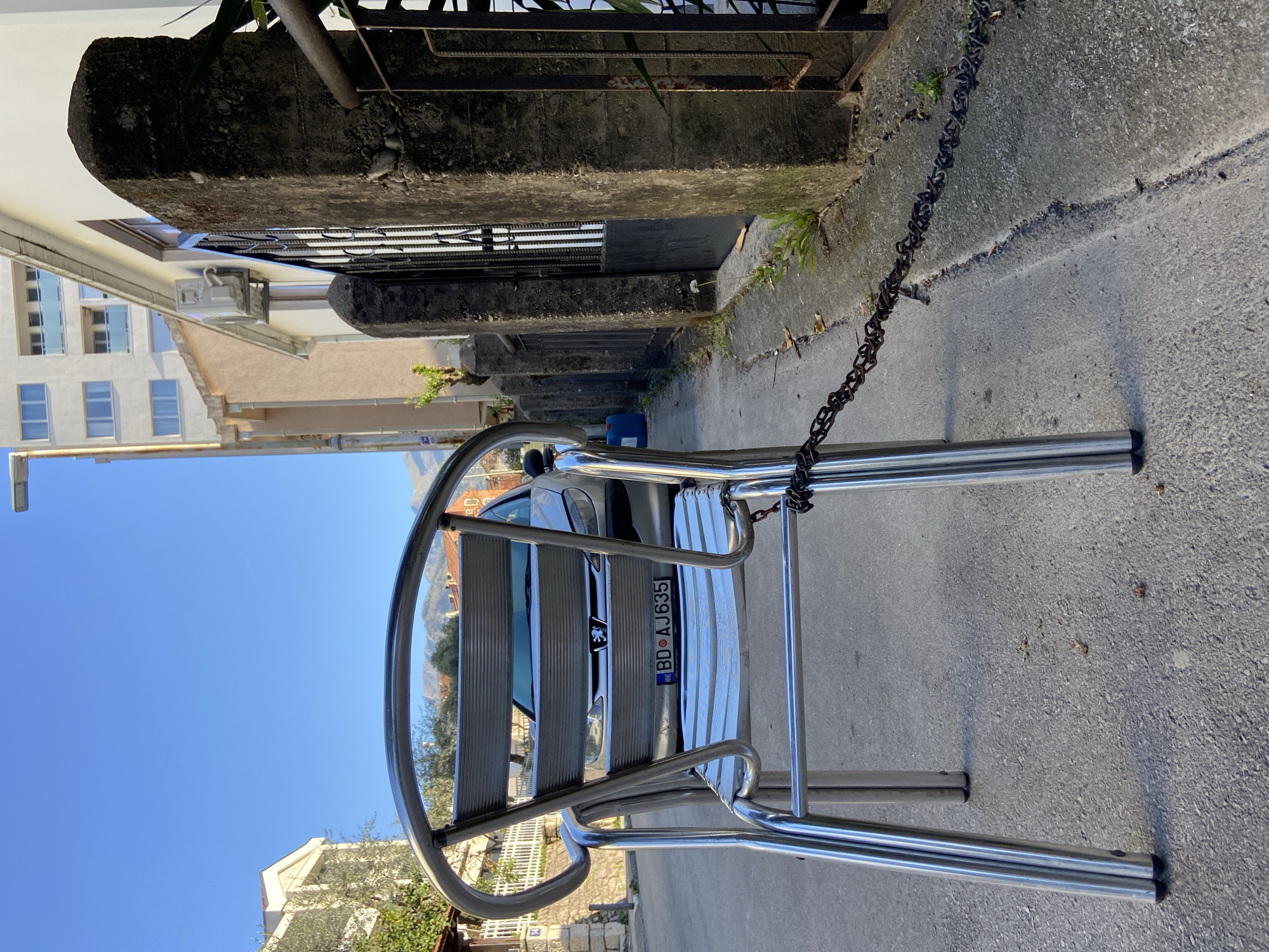 A daytime shot in Budva, Montenegro depicts a chrome metal chair on a gray concrete sidewalk. The chair obscures the rear of a silver car with license plate BD AJ635. A metal chain is attached to the chair. A low, aged stone wall runs along the sidewalk edge, adjacent to a building. In the background, other buildings are visible under a clear blue sky.