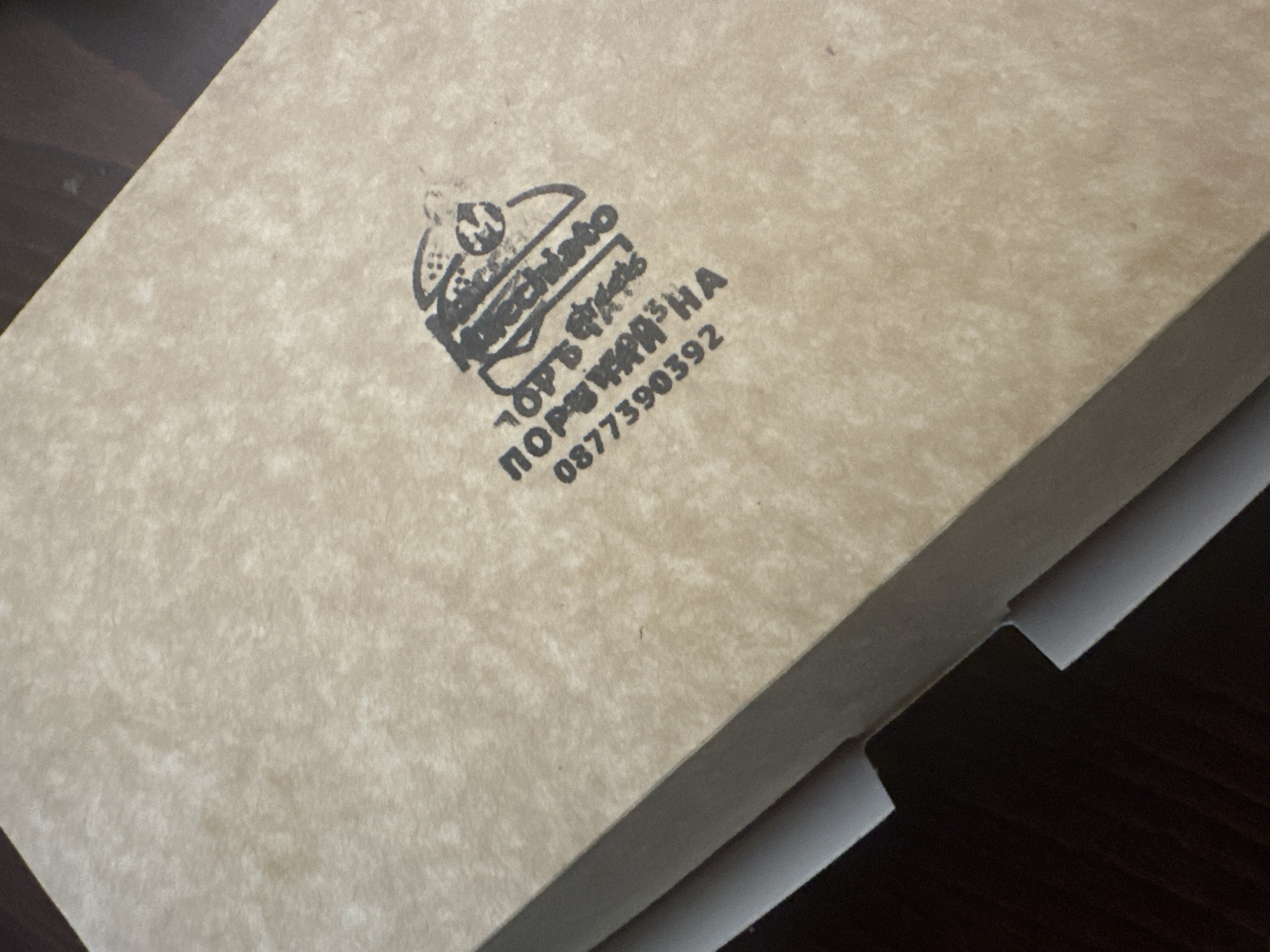 The image shows a cardboard box with a black stamp on it. The stamp includes the word "Kucchiano" in a stylized font, along with some kind of logo above the word. Below the word are two lines of Cyrillic text, "ТОРЪТШАЯЗНА" and then the telephone number "0877390392". The exact location is unknown, but the Cyrillic text suggests the possibility that the location could be in a region using the Cyrillic alphabet.