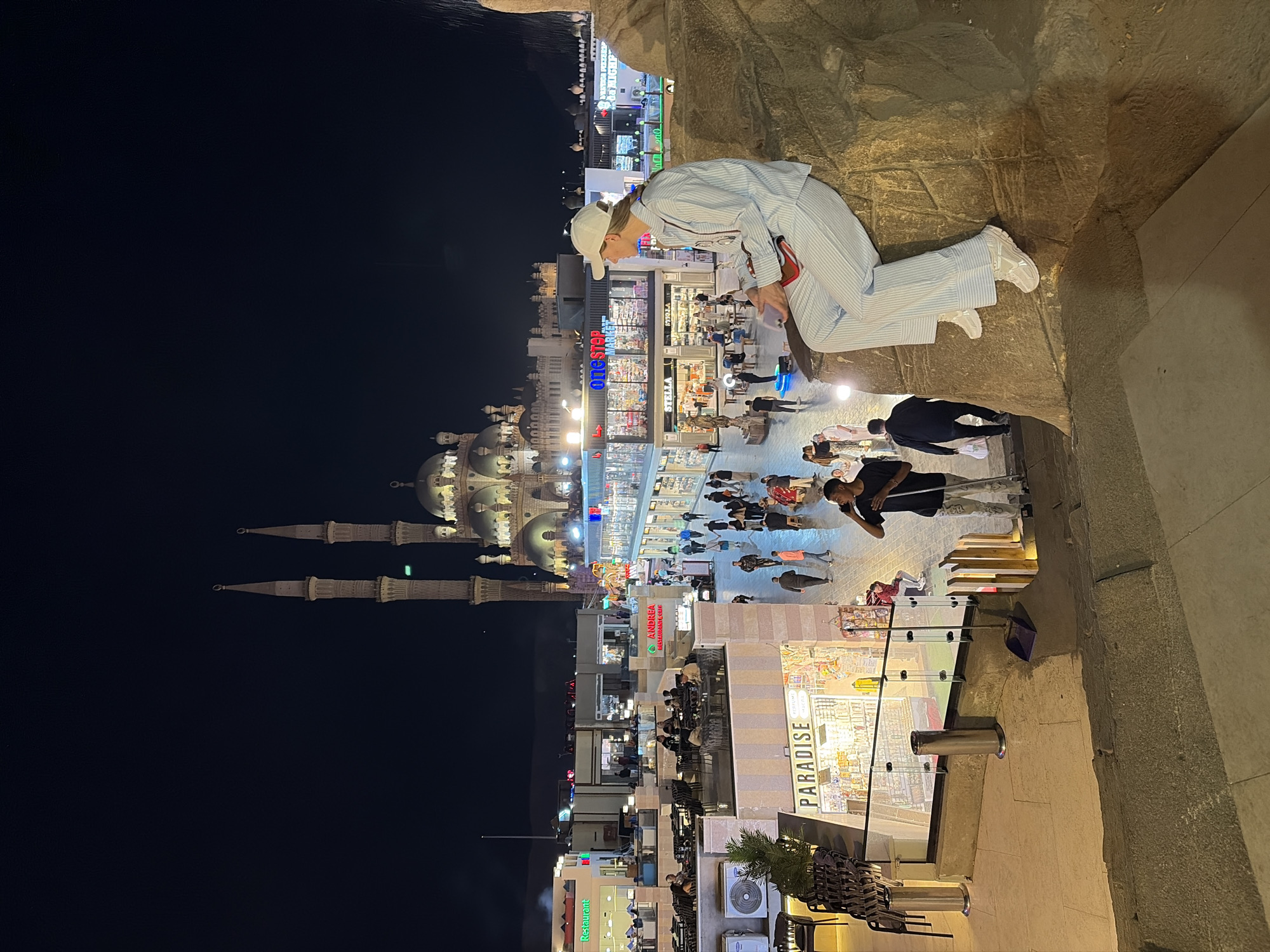 The image depicts a nighttime scene in Sharm El Sheikh, Egypt, specifically within the commercial area leading to the Al Sahaba Mosque. In the immediate foreground, a female individual, wearing a light blue and white striped shirt, matching wide-leg trousers, white sneakers, and a white baseball cap, is seated on a textured, rock-like architectural feature. She holds a small red object, possibly a phone or clutch, in her hands and faces left, observing the scene below.
Below and extending into the midground is a multi-level pedestrian promenade filled with people and shops. Numerous individuals are walking, congregating, or seated at outdoor dining areas. Shopfronts with illuminated signs are visible, including "PARADISE" in the lower left and "One Stop MARKET" and "ANDREA Restaurant" further back. A male individual in a dark t-shirt and patterned trousers is seen in the lower left-center, holding a long stick, possibly a broom, and appearing to clean the ground. Glass railings demarcate the edges of elevated walkways.
Dominating the background is the brightly illuminated Al Sahaba Mosque, featuring a large central dome, multiple smaller domes, and two tall, slender minarets with pointed finials. The mosque is lit with warm white and yellow lights, standing out against the dark night sky. Other commercial buildings and structures surround the mosque. The overall environment is a bustling, artificially lit urban commercial and cultural hub.