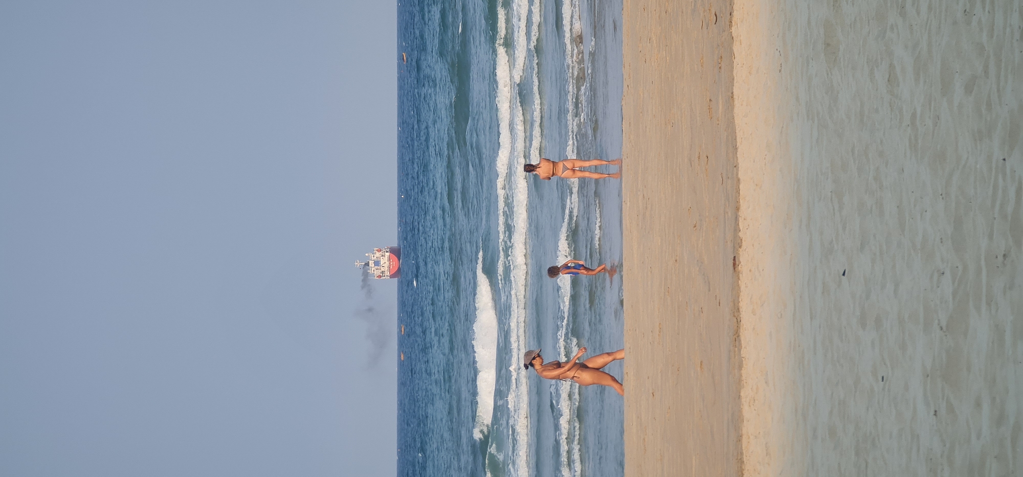 A daytime beach scene captures people enjoying the ocean and sandy shore in Đà Nẵng, Vietnam. The image is composed of a clear, pale blue sky at the top, a vast expanse of blue ocean with white waves in the middle, and a wide strip of light brown sand at the bottom.
In the distance, on the horizon of the ocean, a large red cargo ship is visible, emitting a plume of dark smoke from its funnel as it sails across the water.
Closer to the shore, several individuals are present. One woman, wearing a bikini and a hat, stands on the wet sand at the water's edge, facing away from the camera and slightly to the left, appearing to walk or stand in the gentle surf. Further out in the shallow water, another woman in a bikini is lying face down, partially submerged in the incoming waves, seemingly sunbathing or relaxing. A child or smaller person in a blue swimsuit stands upright in the very shallow water, with their back to the camera. Another person's head and shoulders are visible in the water near the waves, closer to the foreground.
The water is clear, showing the gentle white foamy crests of small waves rolling onto the shore. The sand on the beach is light brown, appearing wet near the water's edge and drier further up. The overall atmosphere suggests a peaceful and leisurely beach day. There is no visible text in the image.