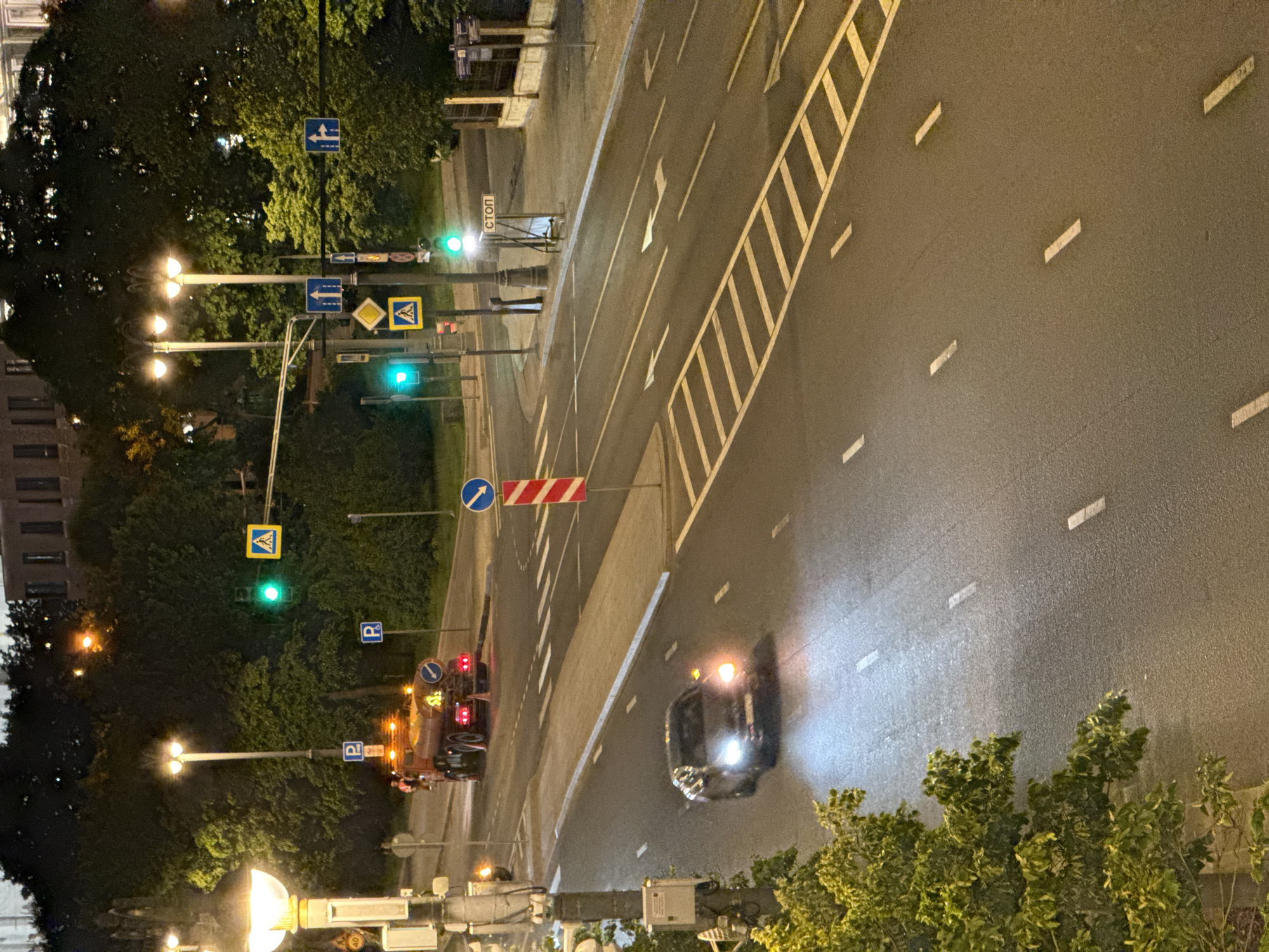This detailed image captures an urban street scene at night in Moscow, Russia, viewed from a slightly elevated position.
The foreground is dominated by a dark asphalt road with white dashed lane markings and solid white lines delineating the shoulder. On the left, a dark-colored sedan, possibly a luxury model, is visible with bright white headlights illuminated and its right amber turn signal flashing. The car is moving away from the viewer. Green foliage from a tree is prominent in the lower left, partially obscuring the view of the road.
The midground features a complex intersection with multiple lanes indicated by painted white arrows on the asphalt, directing traffic straight ahead and right. A hatched pedestrian crossing area is prominently marked with white and yellow stripes across the road. Several lampposts with multiple ornate fixtures illuminate the scene with warm light. Traffic lights are visible, displaying green signals for vehicular traffic. A series of road signs are mounted on poles: a yellow square with a black pedestrian symbol indicating a pedestrian crossing, a blue square with a white "P" for parking, and blue square signs with white arrows indicating permissible directions (straight, right turn). A red and white striped vertical pole stands near the center of the intersection. On the right side of the road, a portable stand with a sign bearing the Cyrillic word "СТОП" (STOP) is visible.
In the background, a large orange/red municipal tanker truck, likely a street-cleaning or watering vehicle, is either stopped or moving slowly on a side road or turn lane. It has several amber flashing lights on its roof and rear, along with red taillights. A person wearing a high-visibility vest is visible near the truck. Behind the intersection and the truck, dense green trees line the street, with dimly lit large buildings of architectural significance visible in the distance through the foliage. Utility poles and various street furniture are interspersed throughout the scene. The sky is dark, indicating nighttime conditions.