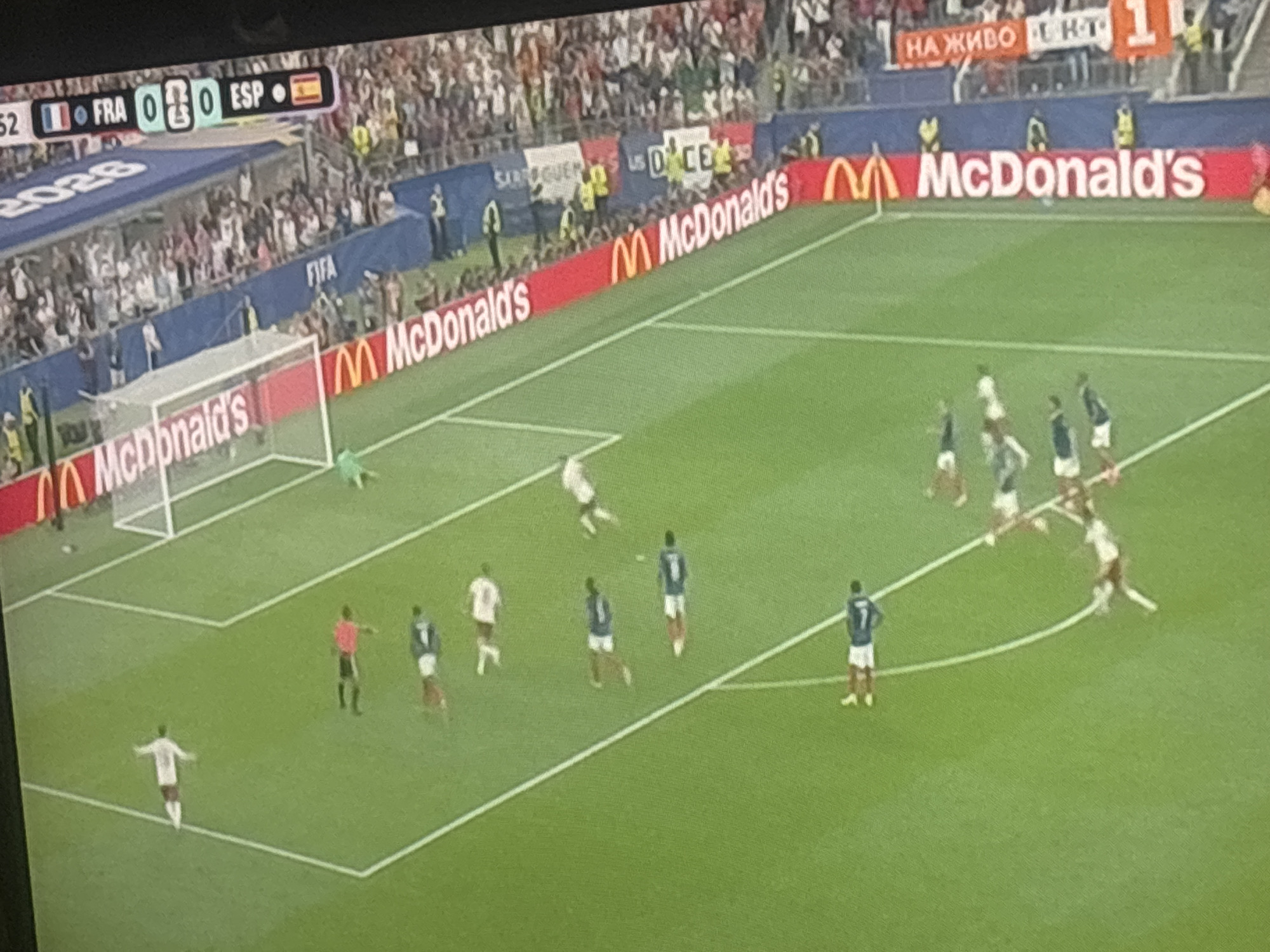 The image shows a football match on a green grass field, with players in white and blue uniforms. There are goalposts and a net visible. Spectators are in the background, seated in stands. A red advertising board with the McDonald's logo is present along the side of the field. In the upper left corner, a scoreboard displays "FRA 0 0 ESP". A banner in the background reads "НА ЖИВО" and "FC". The scene appears to be a professional football game, likely in a stadium. The specific location in Sredets, Bulgaria cannot be determined from the provided image alone, as it lacks any identifying landmarks or text specific to that city.