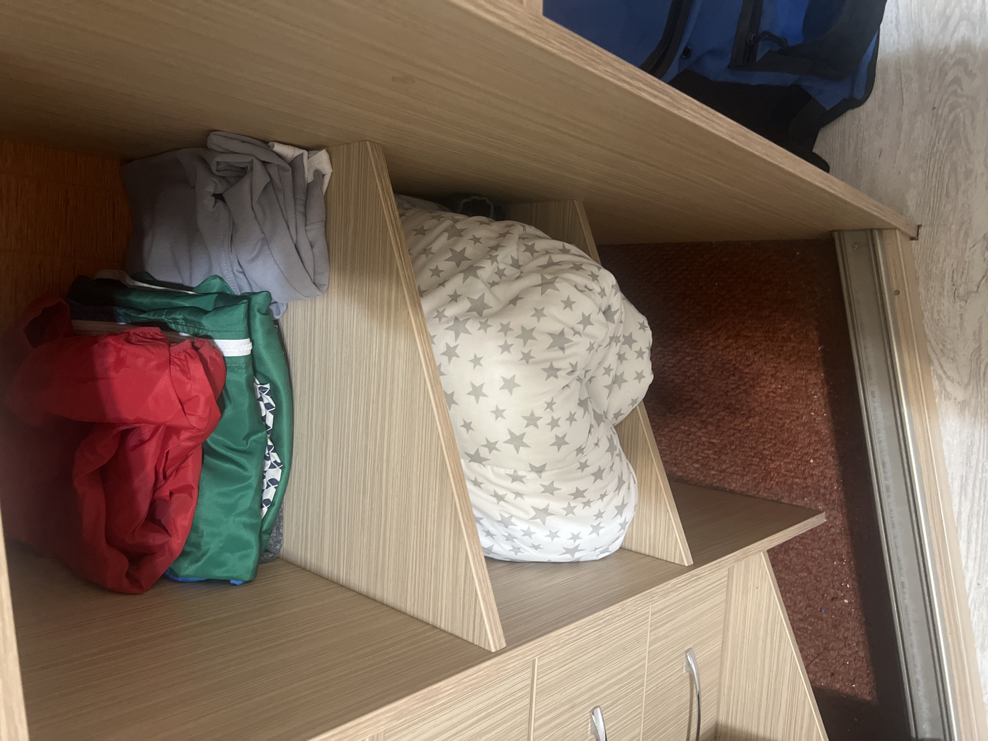 This is a close-up, eye-level shot of the interior of a light-colored wood-grained wardrobe. The wardrobe is divided into shelves. The top shelf contains folded clothing, including a red garment, a green garment, and a grey garment on the right. The middle shelf holds a white pillow with grey stars. The bottom section of the wardrobe shows a reddish-brown carpeted floor and a sliding door track. A blue bag is partially visible to the right of the middle shelf. The scene is static and shows stored items. There are no people or visible actions occurring. The setting appears to be residential. The location is specified as Surgut, Russia.