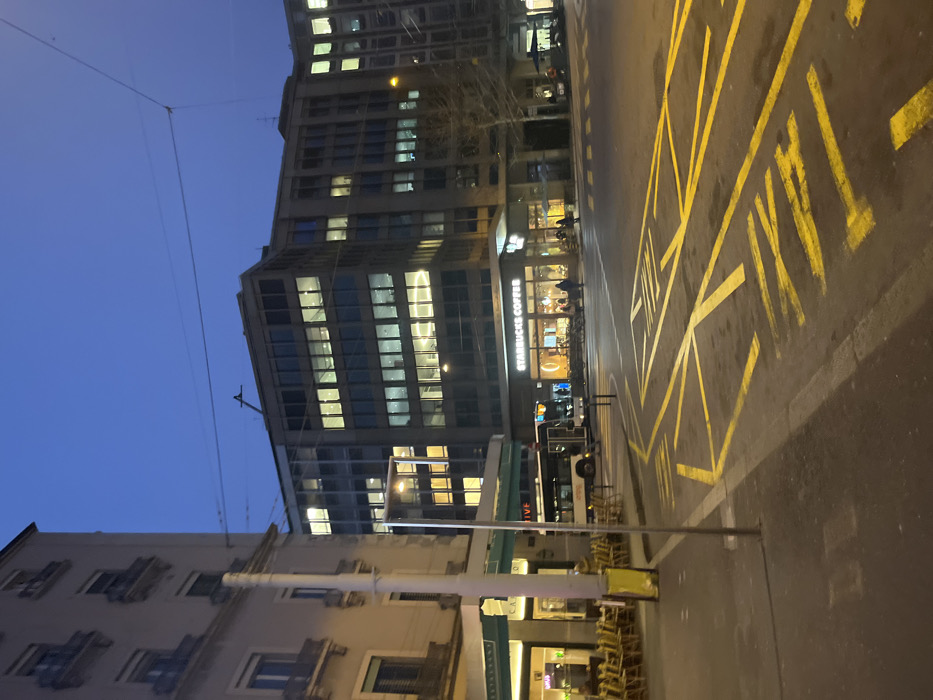 The image captures an urban street scene at dusk or night in Genève, Switzerland. A deep blue sky hangs above a cityscape dominated by buildings and artificial light. On the right, a large modern building, likely an office block, stands tall with many windows brightly illuminated, reflecting light. At its ground level, a "STARBUCKS COFFEE" shop is prominently visible, glowing with interior lights, and a few individuals can be seen inside.
In front of the buildings, a wide paved road is marked with clear yellow lines, including a section with cross-hatching and the word "TAXI" painted on the asphalt, indicating a designated taxi stand. A public transport vehicle, possibly a bus or tram, is partially visible on the street, with the letters "IVE" discernible on its side.
To the left, an older, possibly residential, building with architectural details is partially captured. Overhead utility wires are visible against the dark sky. Below the older building, stacked tables and chairs suggest an outdoor seating area for another establishment. The overall atmosphere is calm and quiet, characteristic of an urban evening.