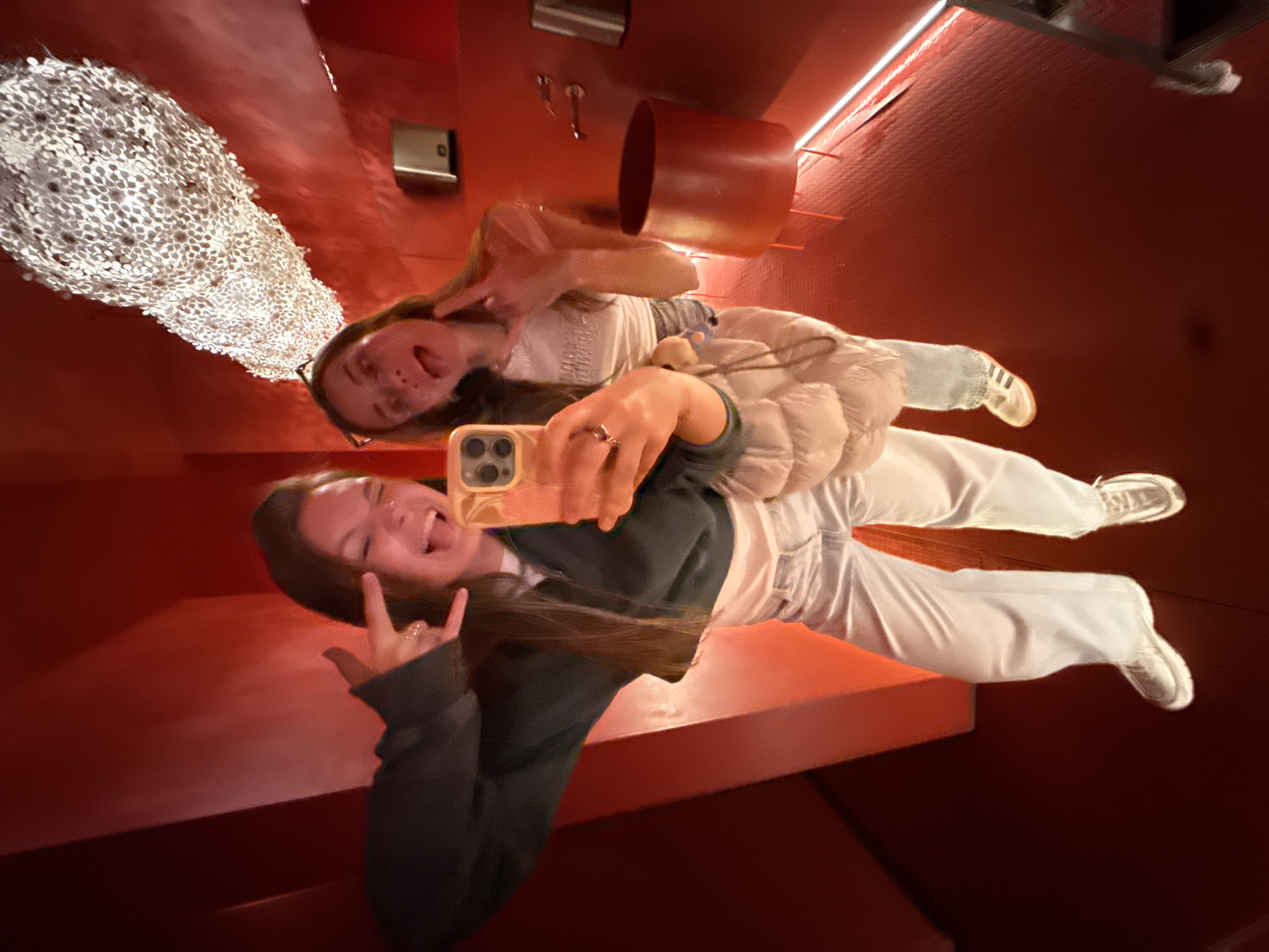 Two young women are captured in a vibrant mirror selfie, likely within a uniquely designed interior space in Belgrade, Serbia. The scene is dominated by a rich, glossy red color that covers the walls and structural elements, giving the impression of an immersive, reflective environment. The image is rotated, presenting a dynamic and slightly disorienting perspective as if the subjects are suspended or leaning on a slanted surface.
In the foreground, one woman holds a light-colored smartphone, capturing the moment. She wears a dark top, light pants, and white sneakers. Her long brown hair frames a wide, joyful smile, with her tongue playfully extended. Her right hand, adorned with a ring, makes a "rock on" gesture. Behind and slightly above her, the second woman is also smiling broadly, making a peace sign with her left hand. She wears glasses, a white shirt, a light puffer jacket, light blue jeans, and striped sneakers.
The background features several distinctive elements against the red surfaces: a large, textured white light fixture or art piece composed of numerous small discs or scales, a metallic rectangular object resembling a vent, a red cylindrical element, and a thin, bright white LED strip running along an edge. The overall atmosphere is lively and cheerful, reflecting a moment of fun between the two individuals in a distinctive, modern setting. There is no visible text.