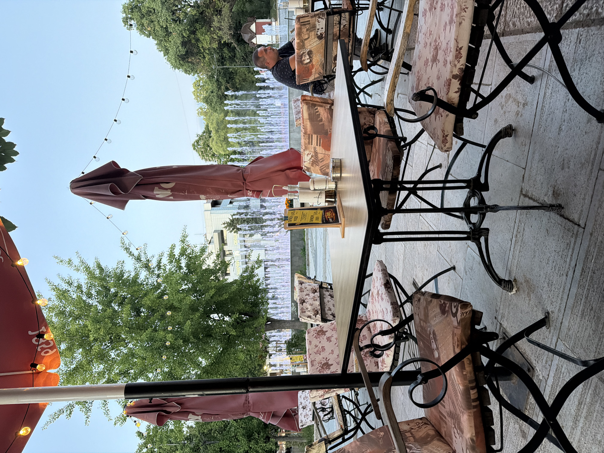 The image depicts an outdoor seating area of a cafe, likely in Svishtov, Bulgaria, given the context. In the foreground, a square wooden table with a dark metal base is set with salt and pepper shakers, a sugar dispenser, and a small menu. Surrounding the table are chairs with ornate black metal frames and cushions patterned with floral and abstract designs. One chair in the foreground is angled towards the viewer, revealing its decorative cushion. In the middle ground, a man with short dark hair is seated at a table, facing to the right and looking away from the camera. He is wearing a dark, patterned t-shirt and appears to be a patron. Behind the seating area, a fountain with multiple spouts of water is visible, illuminated by white and purple lights. Trees with lush green foliage line the background, and beyond them, buildings can be seen. String lights with Edison bulbs are hung across the top of the scene, adding a decorative element to the ambiance. Two large, burgundy patio umbrellas are partially visible, one folded and the other angled. The ground is paved with stone tiles. The overall impression is a relaxed outdoor dining setting, possibly in a public square, with the fountain as a prominent feature.