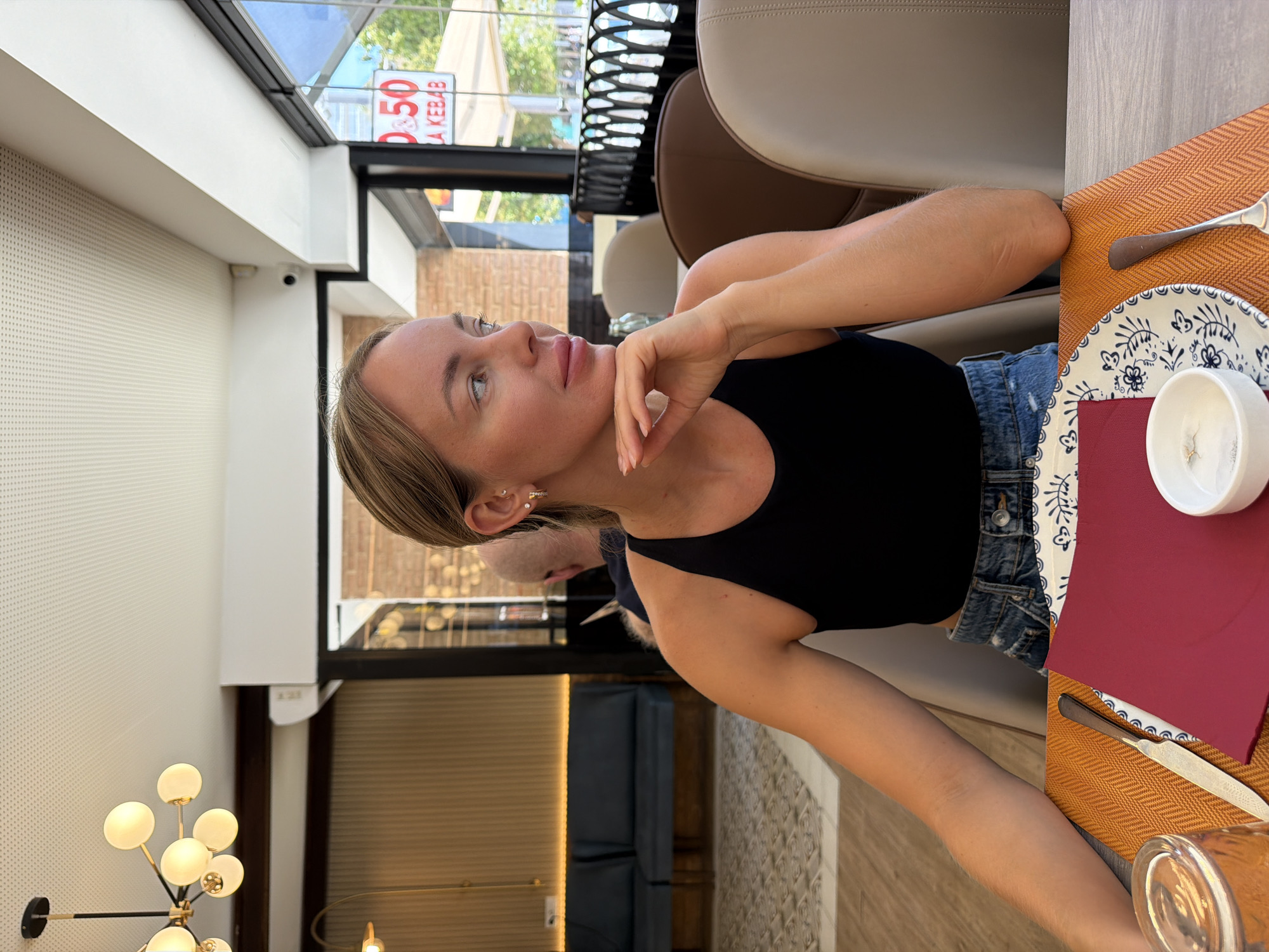 In Benidorm, Spain, a blonde woman in a black tank top and denim shorts sits at a table in an indoor/outdoor restaurant. She is looking to her right, with her chin resting on her hand. On the table in front of her are a red napkin, a plate with a blue and white pattern, a small white bowl, and a glass. The restaurant features a modern chandelier, upholstered seating, and large windows that offer a view of an outdoor seating area and a street. A sign outside reads "0.50 KEBAB."