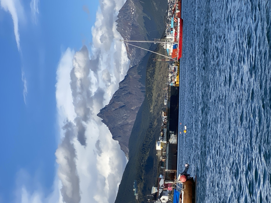 Mountains dominate the landscape, rising behind a coastal town. A ship with a red hull is docked near the town. The sky features blue patches interspersed with white and gray clouds. A body of water occupies the right half of the image, displaying ripple effects on its surface. A portion of a small boat is visible in the lower right corner of the photo.