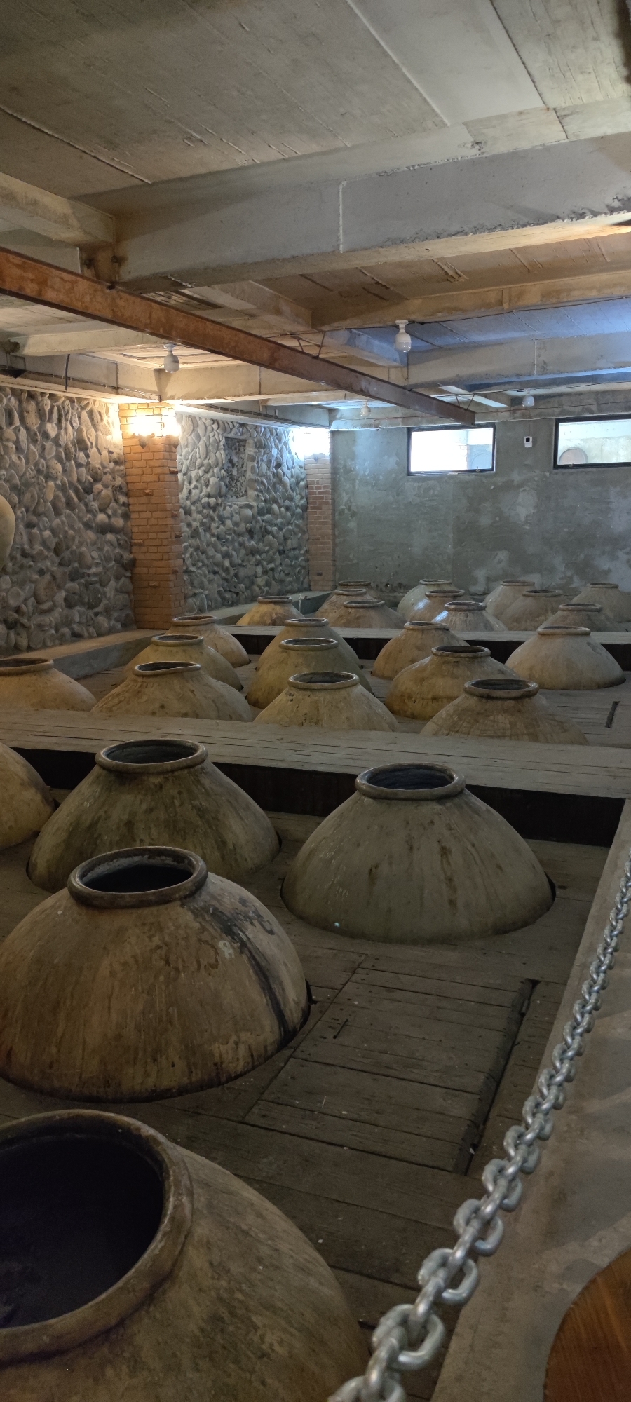 This image depicts the interior of a winery in Voskevaz, Armenia, likely the Voskevaz Winery. The scene is dominated by rows of large, earthenware amphorae (qvevri) partially submerged in wooden troughs on a wooden floor. These qvevri, traditionally used for winemaking in Georgia and Armenia, are conical in shape with wide openings at the top. They appear aged and weathered, some with dark stains.
The environment is industrial, with a concrete ceiling supported by metal beams and wooden structures. Exposed brickwork and a stone wall are visible in the background, adding a rustic texture to the space. Lighting fixtures are mounted on the ceiling. Two small, rectangular windows with dark frames are present on the far wall, offering a glimpse of an exterior. A metal chain runs diagonally across the foreground, separating the viewer from some of the qvevri. No people are present in the image. The overall impression is one of a traditional winemaking facility, highlighting ancient techniques within a functional, albeit somewhat aged, setting.