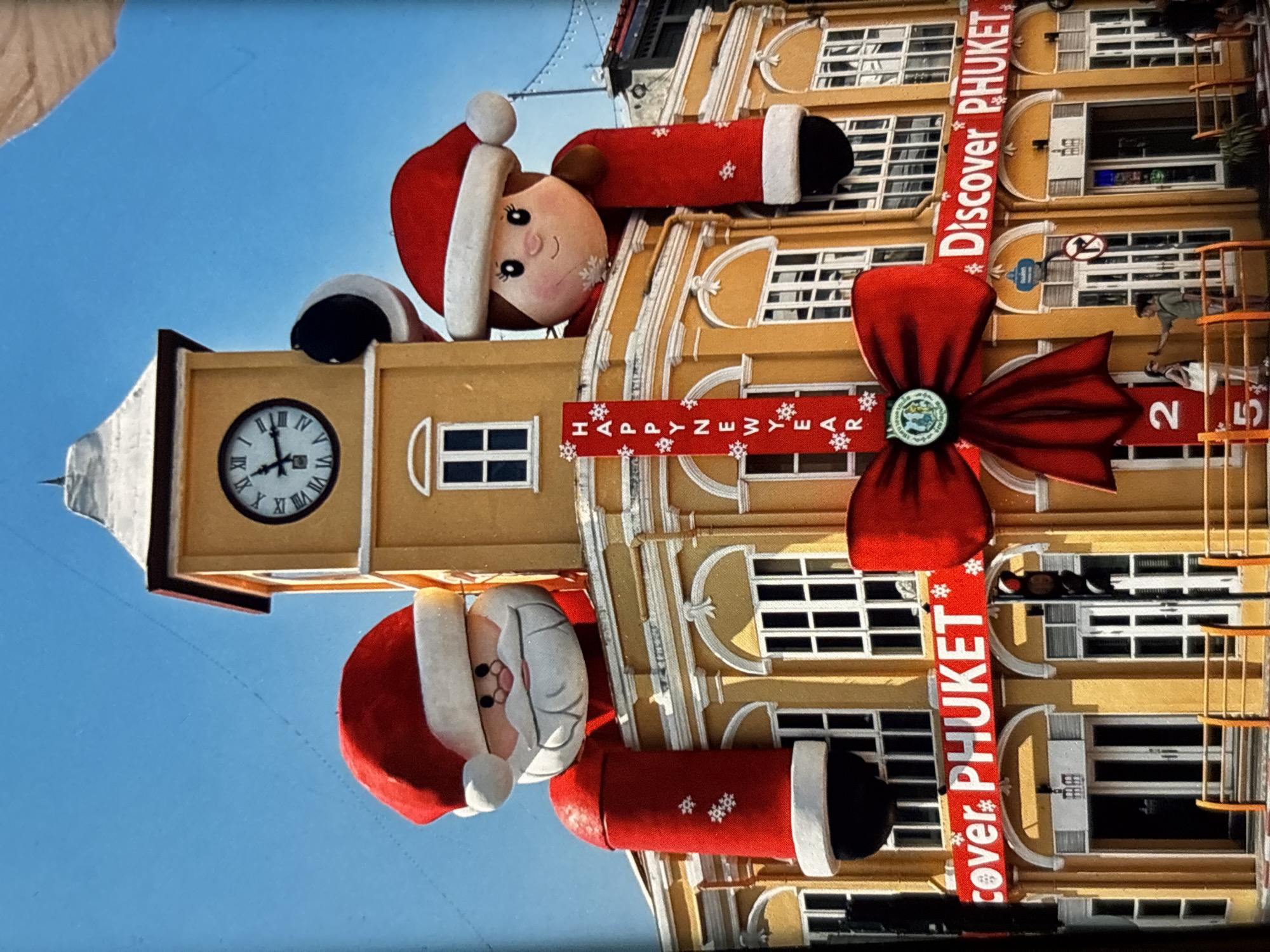 The image depicts the exterior of a yellow building in Chalong, Thailand, adorned with festive Christmas and New Year decorations. Two large inflatable figures, resembling Santa Claus and a female character in Christmas attire, are perched on top of the building, peeking over the edge. A large red bow is prominently displayed in the center of the building's facade, with a red banner beneath it that reads "Happy New Year." Two horizontal banners on the building's lower levels read "Discover PHUKET." A clock tower rises above the building. In front of the building, a traffic light is visible, and a few people are walking by. The sky is clear and blue.