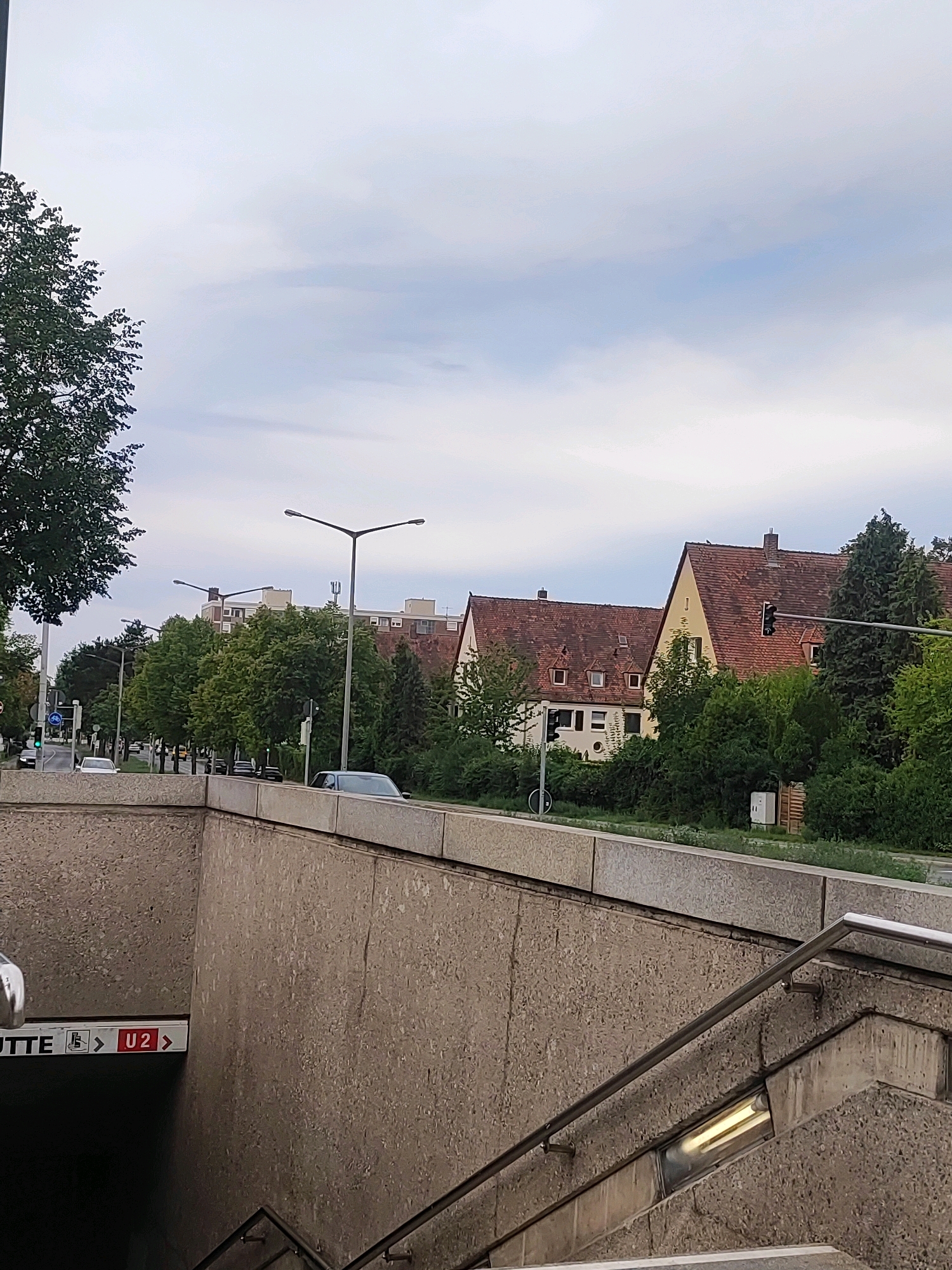The image captures an exterior view of an underground station entrance in Nuremberg, Germany. The station entrance is made of concrete with a metal railing leading down into the darkness. A sign indicates "UTTE" and "U2," suggesting it's a subway station. Above the entrance, a street is visible with cars driving by and trees lining the sidewalk. In the background, there are residential buildings with red-tiled roofs. The sky is overcast with scattered clouds. The overall scene depicts a typical urban environment in Nuremberg on a cloudy day.
