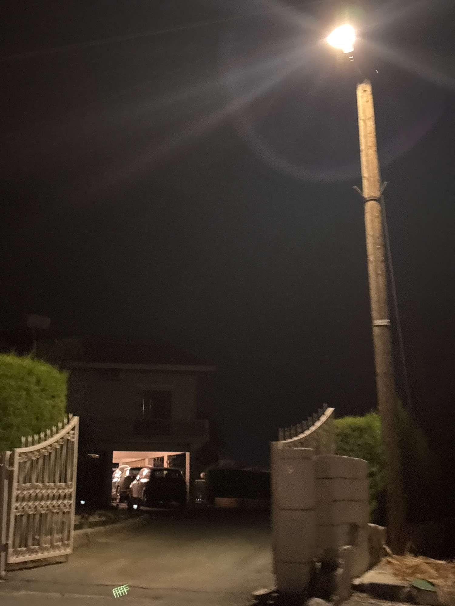 A night time exterior scene in Agios Tychona Community, Cyprus shows a driveway leading to a house with an open gate. A streetlight illuminates the scene from the right. There are cars parked inside the house's carport. A hedge is visible to the left of the gate and a cylindrical concrete structure is to the right of the driveway.