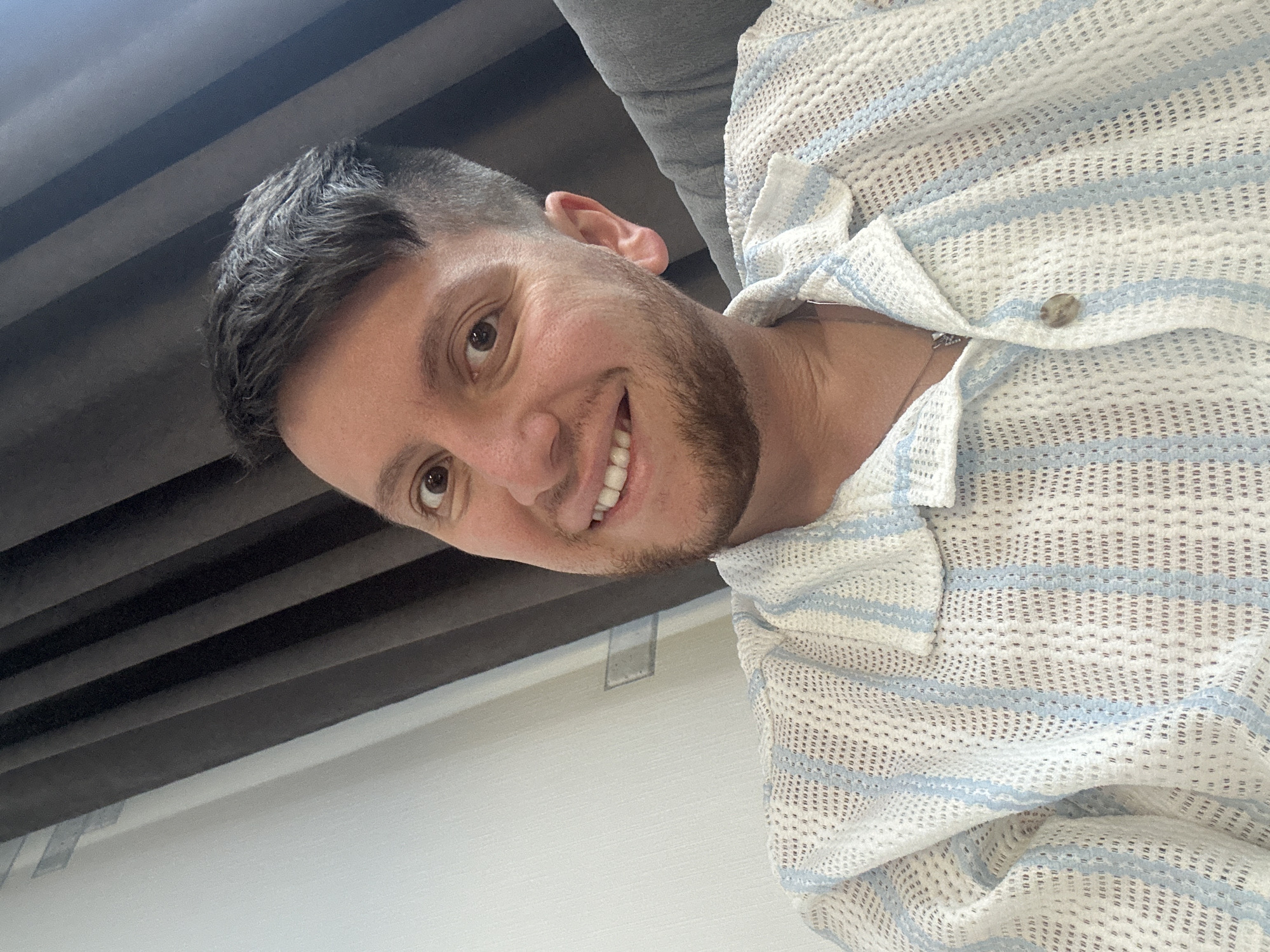 The image shows a close-up of a man's face and upper torso. He has brown eyes, dark hair, and a beard. He is smiling and wearing a white, short-sleeved shirt with blue stripes. The background is blurred and appears to be a room with dark curtains on the left and a grey upholstered chair on the right. There is a white wall to the left of the man. The location is identified as Мирный, Russia.