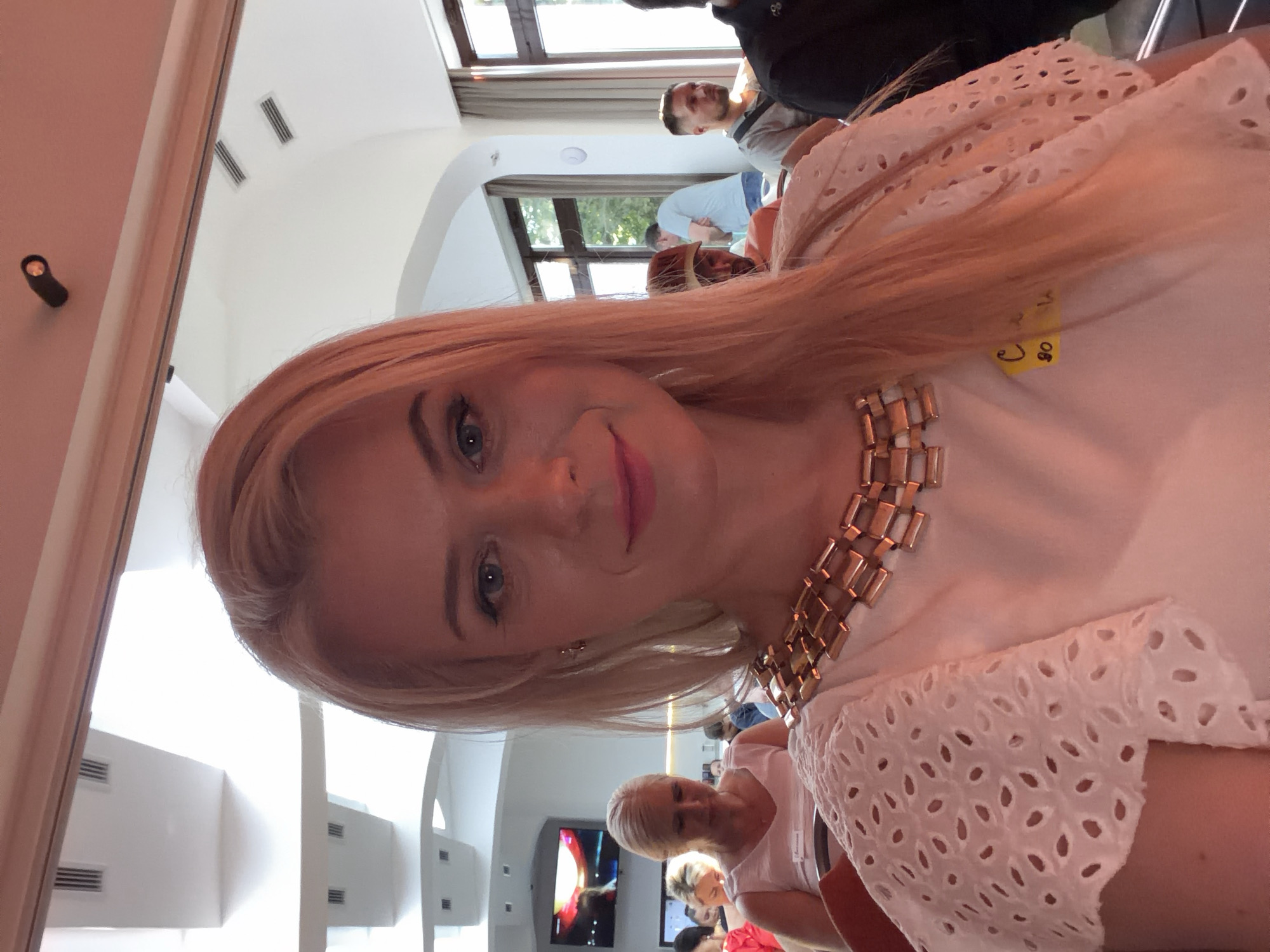 The image is a selfie of a blonde woman with blue eyes wearing a white top with cut-out details on the sleeves and a gold necklace. She is at an indoor event, possibly a conference or seminar, in Sochi, Russia. In the background, other attendees can be seen sitting at tables. There is a screen displaying an image in the far left background. Natural light is coming from windows on the right side of the frame. A yellow nametag is visible on the woman's shirt.