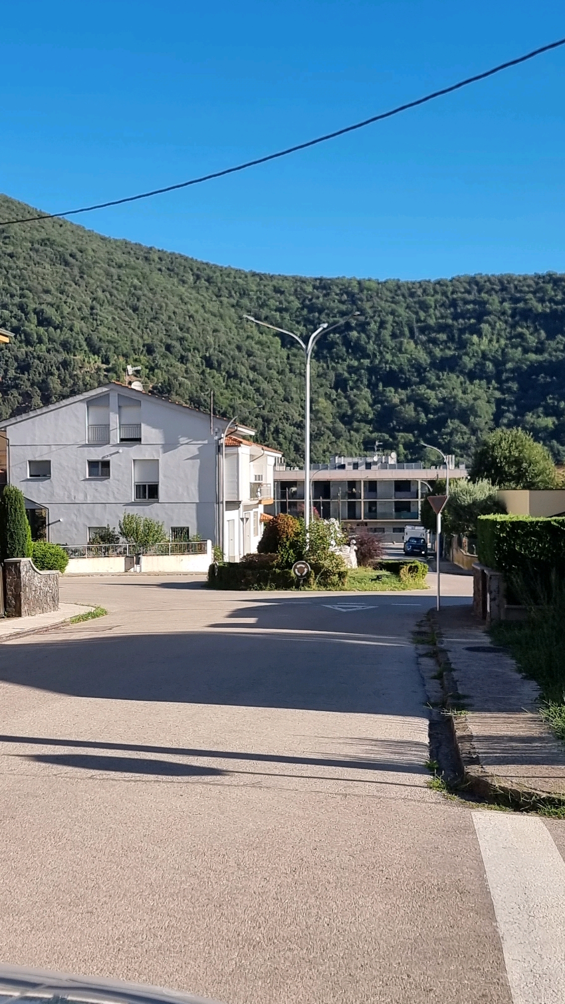 The image captures a street scene in Sant Joan Les Fonts, Spain, under a clear blue sky. A light gray, multi-story building with visible windows and balconies is situated on the left side of the frame. Adjacent to it is a smaller white building with a red-tiled roof. Across the street, a larger, modern building with multiple floors and windows is visible. A street light stands prominently in the center of the image. The road in the foreground casts long shadows from an unseen object, suggesting it is a sunny day. Green foliage and trees surround the buildings, and a densely wooded hillside forms the backdrop. A white pedestrian crossing is marked on the road in the lower right corner. A dark-colored vehicle is parked in front of the modern building. The scene appears quiet and residential.