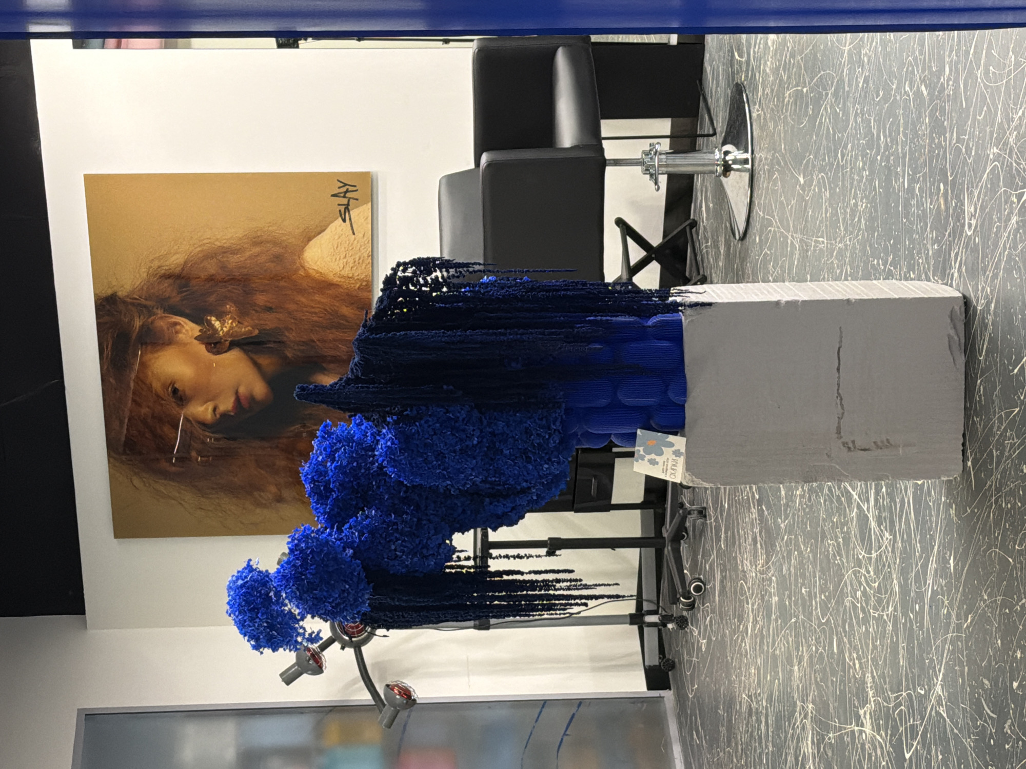 The image shows a creative arrangement of blue artificial flowers and dark blue yarn on a pedestal in what appears to be a hair salon in Saint Petersburg, Russia. The backdrop includes a framed image of a woman with flowing hair and distinctive earrings, as well as salon chairs and equipment. The floor is a dark color with white paint splatters. The overall aesthetic suggests a modern and artistic salon environment.