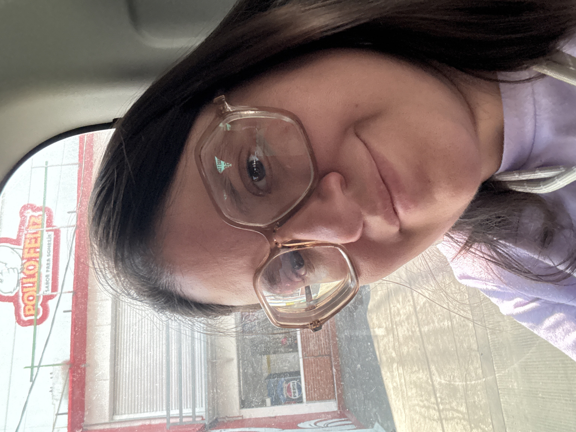 A selfie taken from inside a car shows a person wearing eyeglasses and a light purple hoodie, facing towards the camera. The background seen through the car window shows a building with the words "POLLO FELIZ" prominently displayed, indicating the location is likely near a Pollo Feliz restaurant. The building is characterized by a red and white color scheme. The presence of a metal roller shutter suggests the building could be a commercial establishment. The photo appears to have been taken in Jiutepec, Mexico, based on the presence of a Pollo Feliz restaurant, which is a common establishment in Mexico.