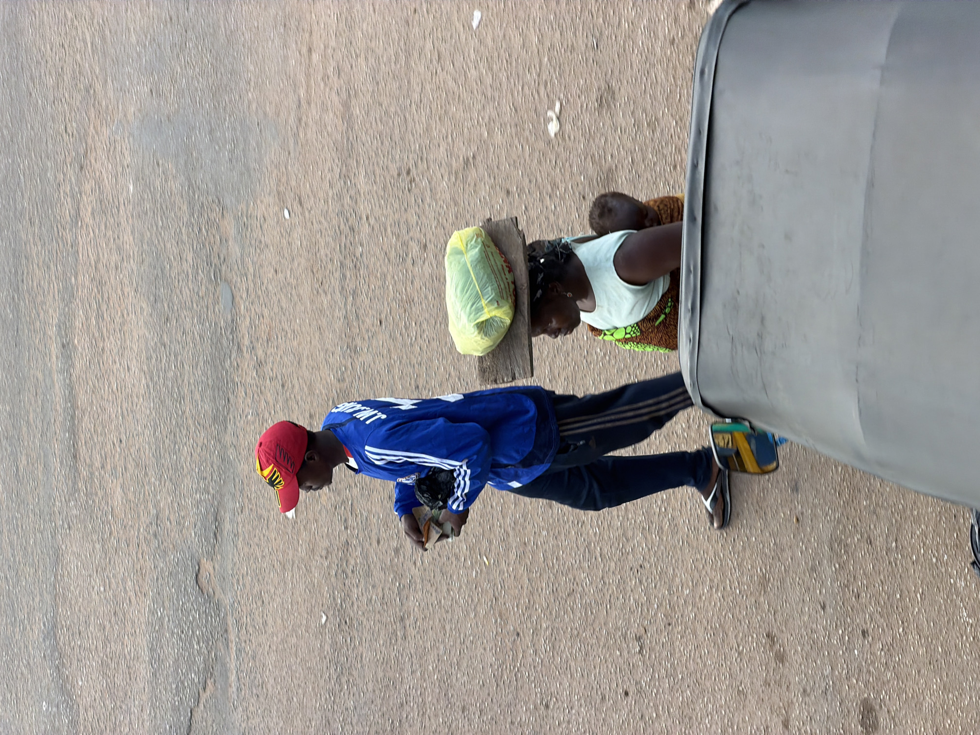 In Bukuru, Nigeria, a man wearing a red and yellow cap and a blue long-sleeved shirt with white stripes on the sleeves walks across a textured, tan-colored asphalt road. He is carrying a small black object in his left hand and what appears to be money or receipts in his right hand. To his right, a woman balances a large, light green, plastic-wrapped object on a wooden plank on her head. She is wearing a light blue sleeveless top and a patterned wrap around her lower body. A young child is strapped to her back, also wrapped in the same patterned fabric. In the foreground, the hood of a dark-colored vehicle, possibly a tricycle or a small truck, occupies the bottom portion of the image, its textured surface reflecting the light. The scene captures a moment of daily life on a Nigerian street.