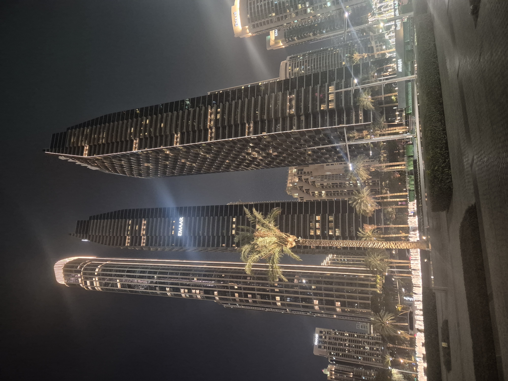 Here's a description of the image:
The image is a night scene in Dubai, United Arab Emirates. It shows several tall buildings illuminated at night. In the center of the frame are three prominent skyscrapers, their facades marked by long vertical windows. One of the towers displays the "EMAAR" logo. Palm trees are interspersed between the buildings, with streetlights providing additional illumination along the road. The lighting emphasizes the buildings' modern architectural details. The ground level appears to be a paved area or sidewalk, slightly blurred. The sky is dark, creating a high contrast with the brightly lit buildings.