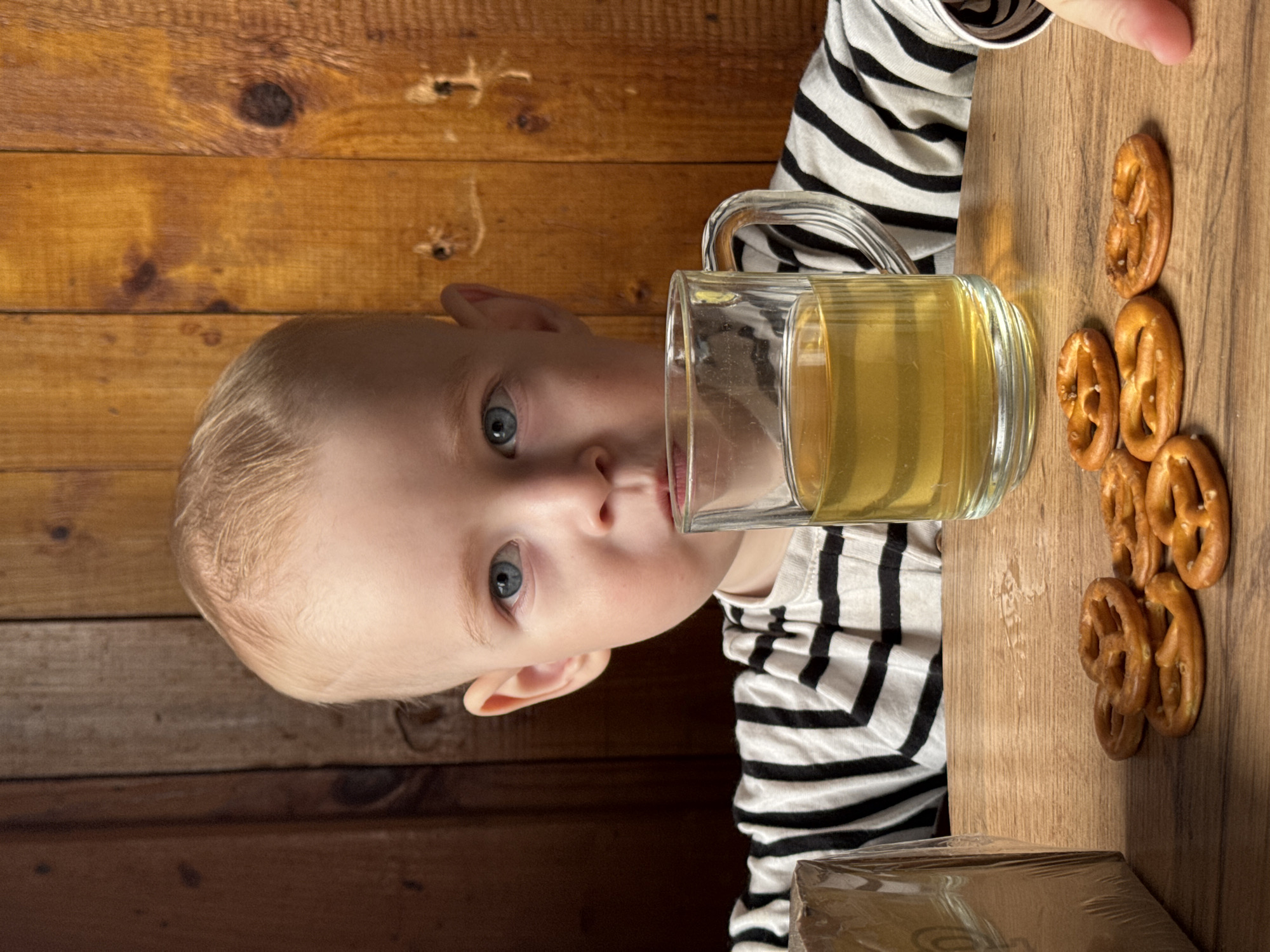 A child is drinking from a clear glass mug filled with a yellow liquid. The child has fair skin, blond hair, and blue eyes, and is wearing a striped black and white shirt. To the right of the mug are several pretzels arranged on a light brown wooden surface that appears to be a table. The background features a wooden wall with a knotty grain. A package partially visible in the bottom left corner is covered in clear plastic. The location is Уральское, Russia.