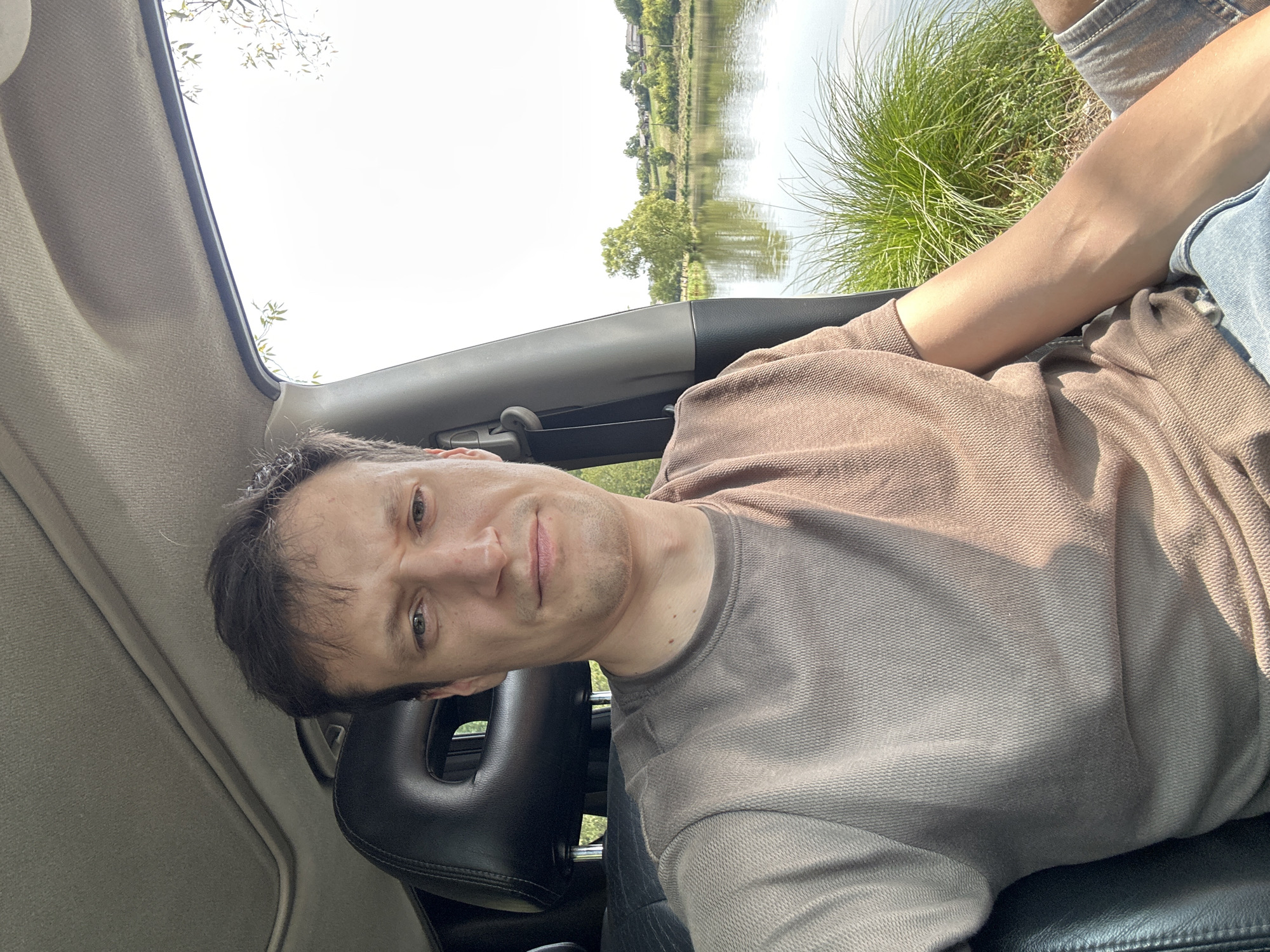 A medium shot shows a man sitting in the driver's seat of a car, facing the camera. The man has brown hair and is wearing a light brown t-shirt. His left arm is resting on his lap, and his right arm is visible on the right side of the frame. The interior of the car, including the car's headrest and ceiling, is visible. Outside the car, a lake is visible with green grass and trees on the opposite shore. The sky is bright and overcast. The image appears to be taken in Makiv, Ukraine.