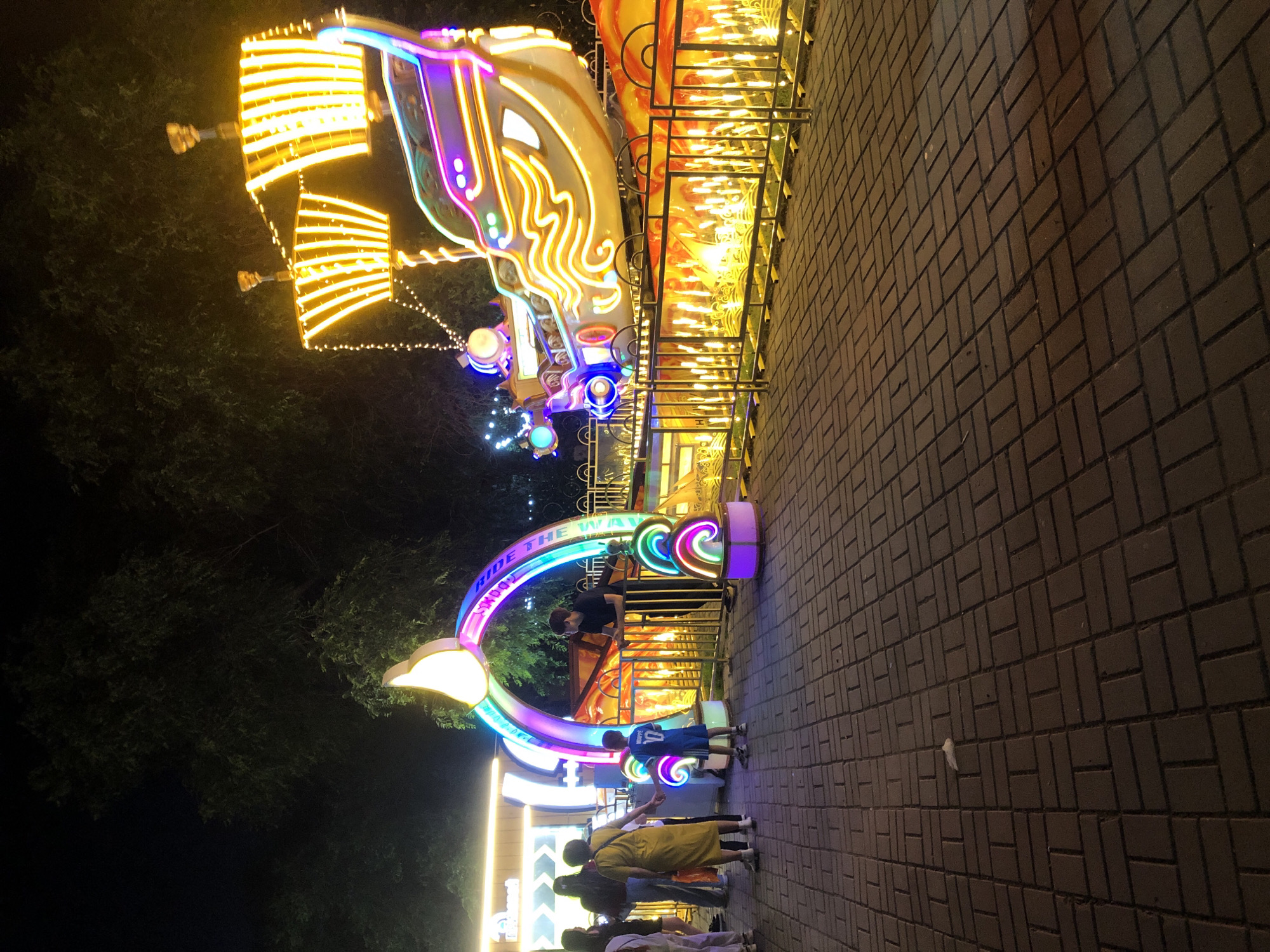An outdoor scene at night depicts a festival or amusement park with a focus on illuminated attractions. In the foreground, a paved walkway made of dark rectangular bricks leads the viewer into the scene. To the left, a group of people, including adults and children, are gathered near a large, brightly colored archway adorned with rainbow-like neon lights. One child, wearing a blue shirt with the number 10 and the name "Mbappe" on the back, stands close to the arch. Adjacent to the arch is a white structure with a canopy, possibly a ticket booth or entrance.
The main attraction in the mid-ground is a large, elaborate illuminated sculpture of a pirate ship. The ship is outlined in vibrant neon lights of yellow, purple, and blue, with its sails depicted as glowing yellow stripes. The ship appears to be a ride or a static display. Surrounding the ship is a fence with numerous small, bright yellow lights that create a fiery effect. In the background, dark trees are visible against the night sky, with some scattered string lights further enhancing the festive atmosphere. The overall impression is one of a lively, colorful nighttime attraction.