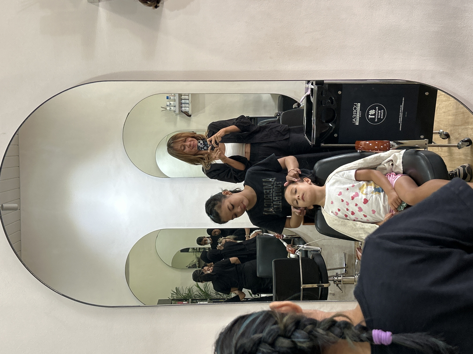 In a bright, modern hair salon, a young girl with closed eyes sits in a black salon chair, her head tilted back slightly as a woman, also in a black t-shirt, gently massages her temples. The girl wears a white t-shirt adorned with a pattern of pink hearts and a cartoon character, and pink shorts. A light-colored cardigan is draped over her shoulders. The salon features large, arched mirrors that reflect the various individuals and the space. In the foreground, the back of another person's head is visible, with their dark hair styled in a prominent braid. Behind the primary interaction, in the reflection of a large mirror, another woman is taking a selfie with her phone. She wears a white crop top and a dark, oversized button-up shirt, with dark pants. Other individuals, also appearing to be salon staff in black attire, are visible in the background reflections, some wearing face masks, and a shelf with various hair products can be seen. The salon appears to be located in North Kuta, Indonesia, given the context provided by the city.