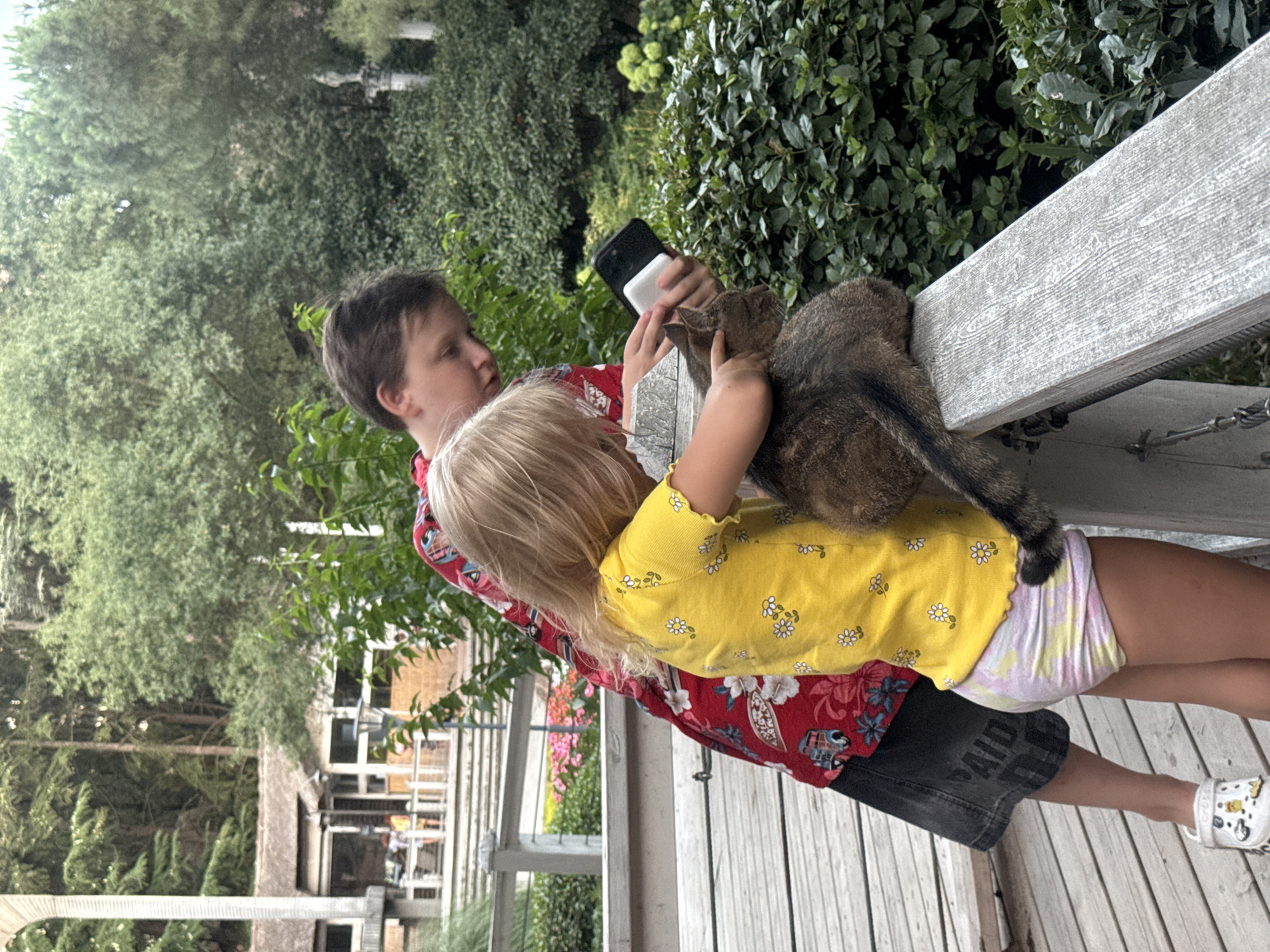 A young girl and boy are interacting with a cat on a wooden deck. The girl, wearing a yellow floral shirt and tie-dye shorts, is petting the cat, which is sitting on a wooden railing. The boy, wearing a red floral shirt and black shorts, is holding a smartphone and a white object, possibly a portable charger. The background shows lush green trees and foliage, suggesting an outdoor setting. The image does not contain any information about the city of Yalta, Ukraine.