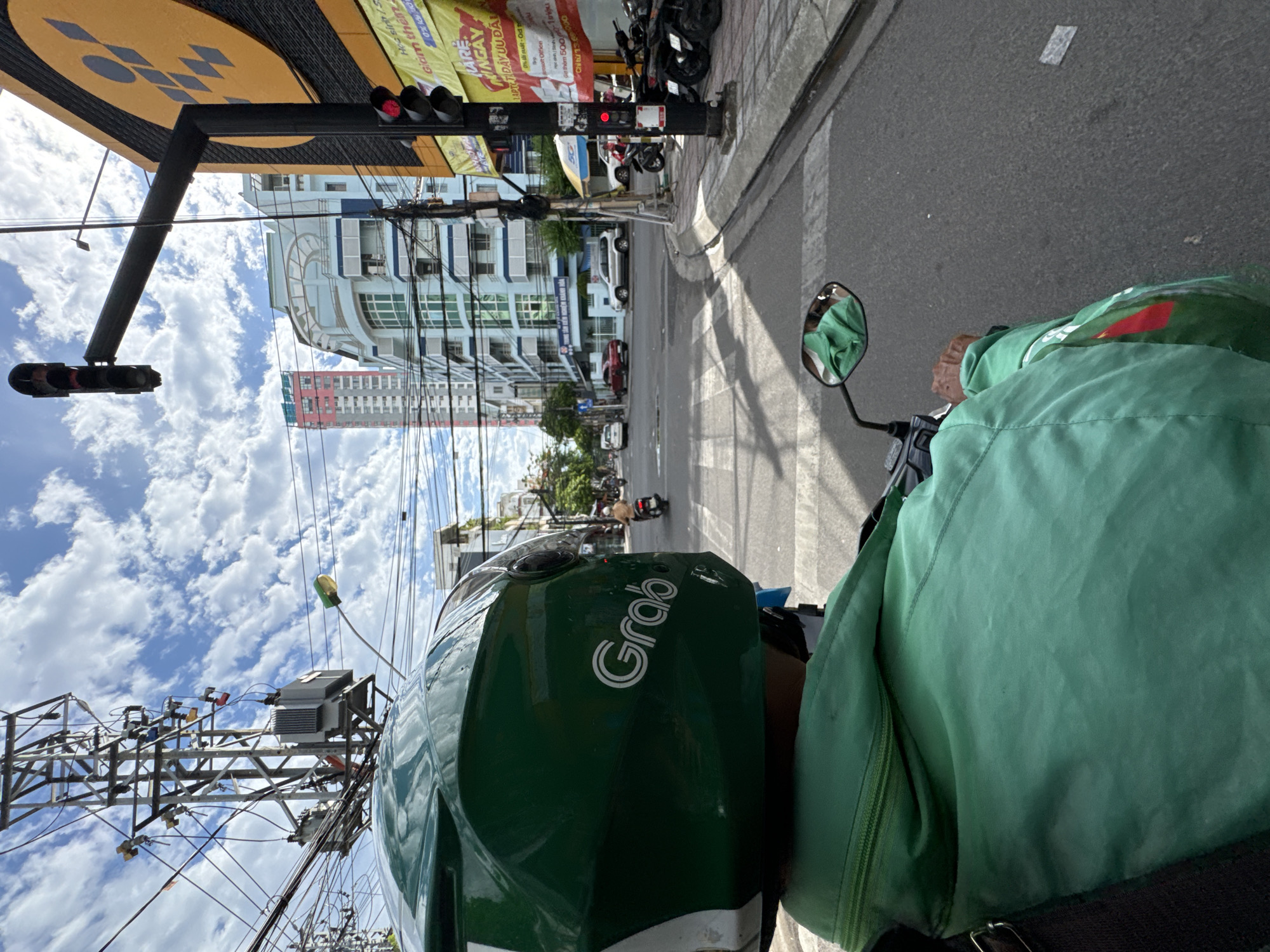 The image depicts a first-person view from a motorcycle in Nha Trang, Vietnam. The rider, wearing a green Grab helmet and jacket, is approaching an intersection. In the foreground, the green helmet with the white "Grab" logo is prominent. The rider's green jacket is also visible. The street is paved asphalt with white pedestrian crossing lines.
In the background, traffic lights are visible, with one red light illuminated on a traffic signal pole to the right. Several motorcycles are parked on the sidewalk. Buildings line the street, including a tall, slender pink and white building, a light blue building with rounded architectural features, and a yellow building with a circular graphic. Power lines and utility poles are abundant, characteristic of urban Vietnamese streetscapes. The sky is partly cloudy with a bright blue expanse.
The intersection appears to be a busy urban area with ongoing traffic, indicated by other vehicles, including cars and motorcycles, in the distance. The scene captures a typical street view in Nha Trang, characterized by its motorcycles, vibrant urban architecture, and overhead infrastructure.