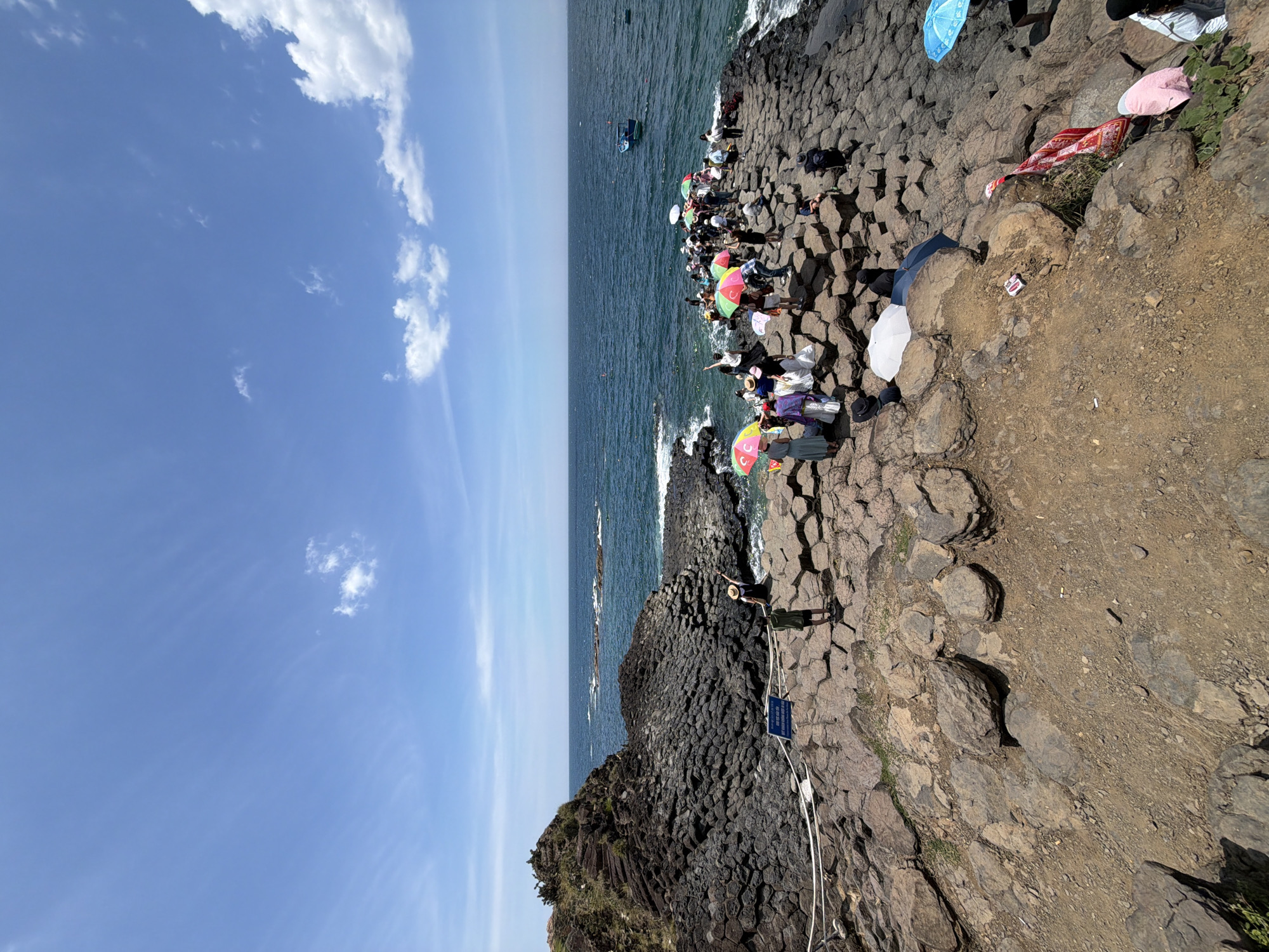 The image depicts a rocky coastal landscape with a clear blue sky and scattered white clouds. The foreground is dominated by a dark, hexagonal rock formation that extends into the ocean. A group of people are gathered on the rocks, many holding colorful umbrellas for shade. Some individuals are standing and observing the scenery, while others are seated. The sea is a deep blue, with a few small boats visible in the distance. The overall environment suggests a popular tourist destination, possibly within Phú Lương, Vietnam, known for its unique geological features.
