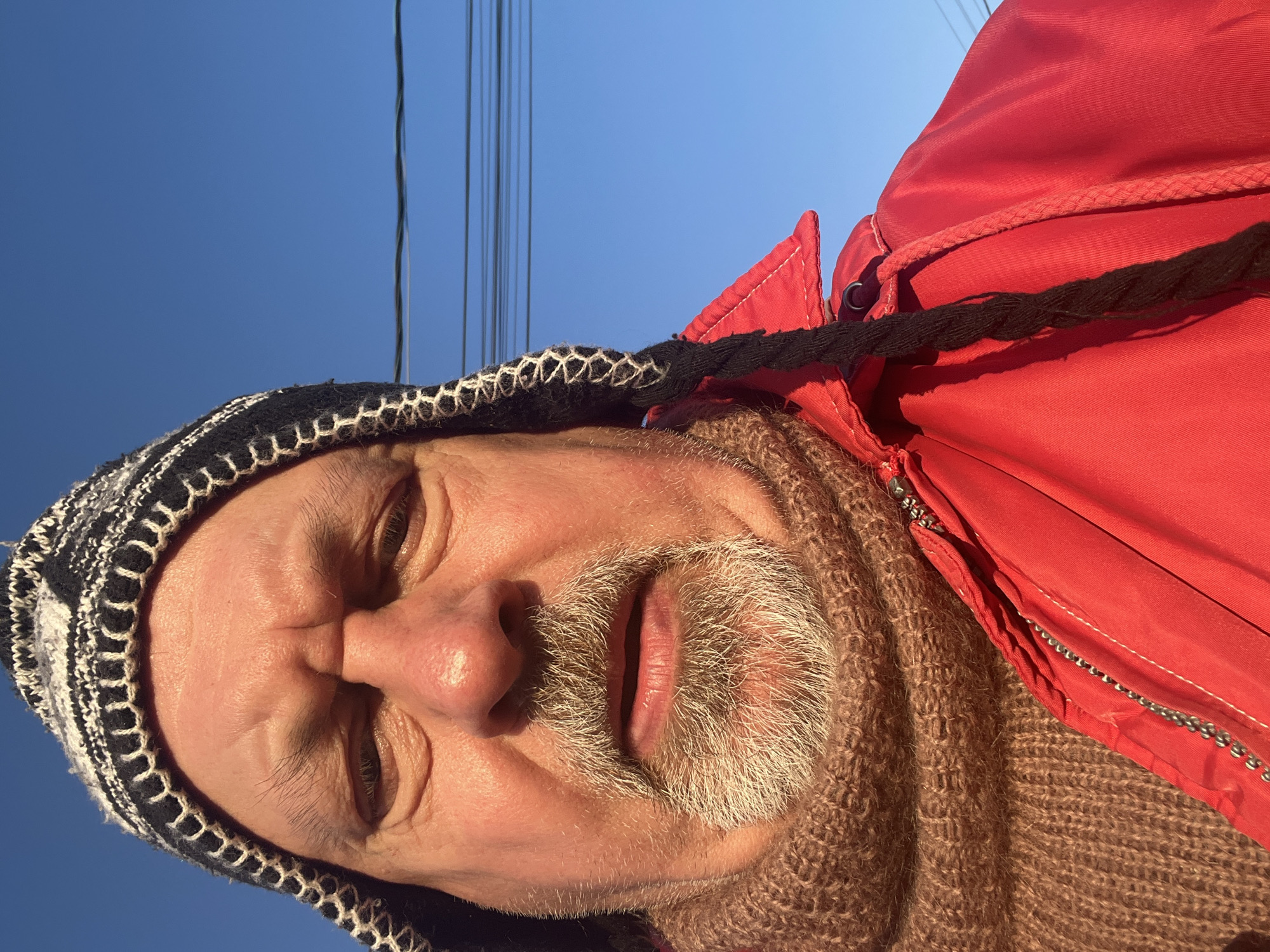 A close-up, low-angle shot depicts a fair-skinned, middle-aged man with a partially grey beard and a knit hat. He is wearing a brown scarf and a red jacket. Overhead power lines are visible against a clear blue sky. The location is not identifiable as Podolsk, Russia, from the given image.