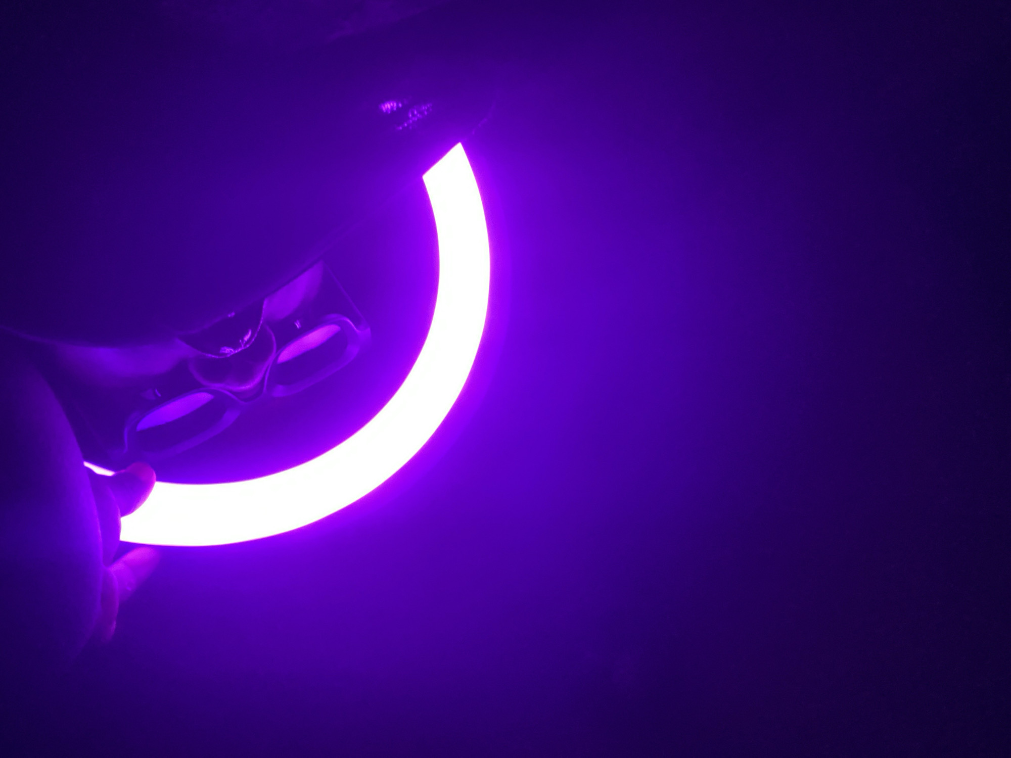 The image captures a close-up view of a hand holding a glowing, curved light source in a dark, purple-lit environment. The main subject is a segment of a bright white, crescent-shaped light, likely a ring light or similar LED fixture. This light emanates a strong purple glow that illuminates the surrounding darkness, creating a vibrant, monochromatic atmosphere.
A human hand, visible in the bottom left corner, gently holds the dark casing of the light fixture. The fingers are curved around the device, suggesting it is being held or positioned. The dark casing of the light device itself appears to be made of plastic or metal, and its surface shows subtle reflections from the bright light.
The scene takes place indoors, in what appears to be a darkened room or studio, designed to highlight the light source. There are no specific location cues to identify Maigizo, Nigeria, or any other external environment. The background is uniformly dark purple, a result of the light spill, with no other objects or details visible. The time of day cannot be determined, but the artificial lighting implies an indoor setting where natural light is absent or blocked. No activity beyond holding the light is clearly depicted, and no specific emotions are discernible. There is no visible text in the image.