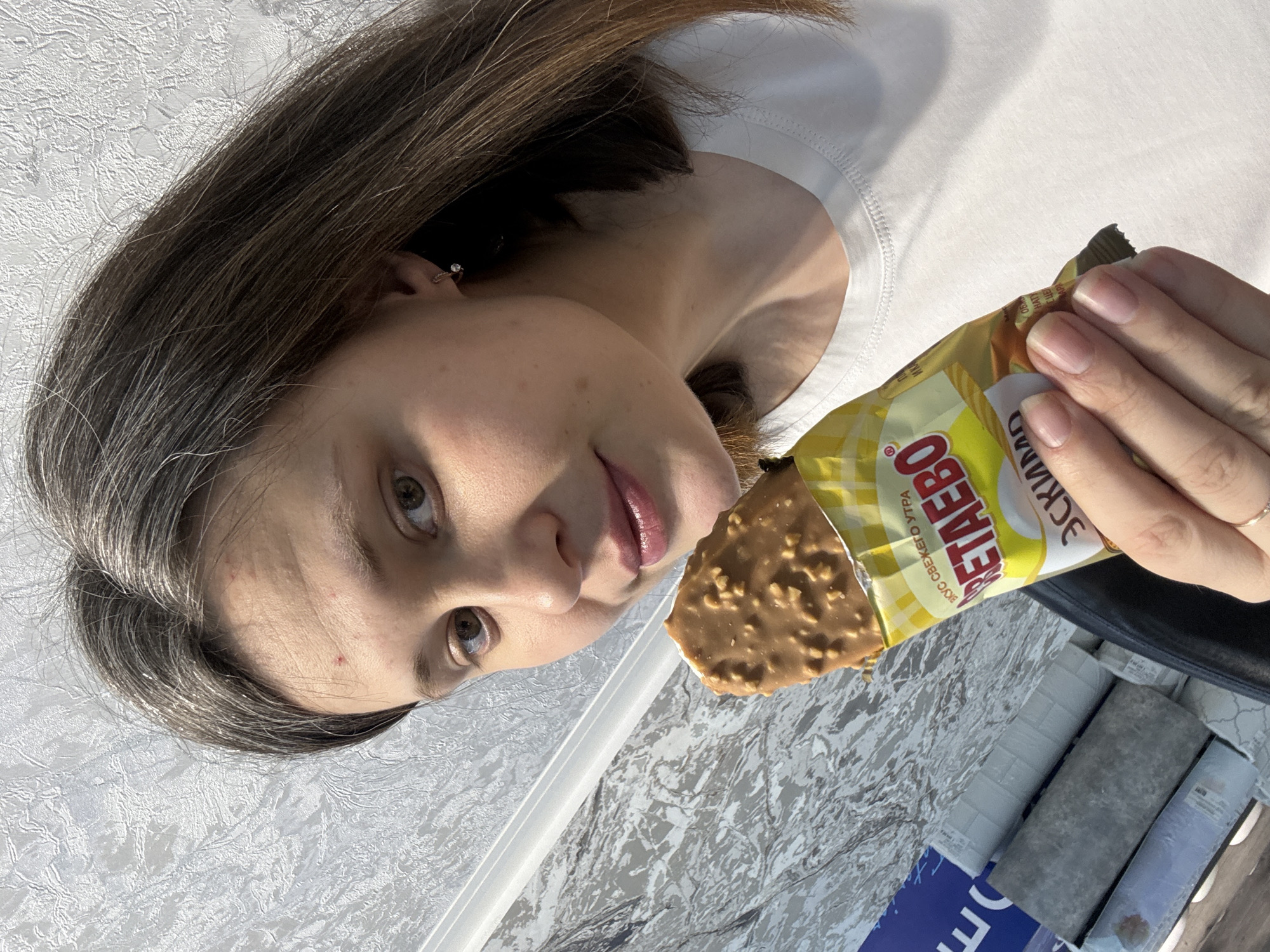 A woman is holding an opened chocolate bar in front of her. The packaging of the chocolate bar is yellow and white. The woman has brown hair with strands of gray. She is wearing a white t-shirt. The background appears to be a room with patterned wallpaper. Rolls of wallpaper are visible in the lower right corner of the image. The brand name on the packaging is 'RETAEBO' written in Russian. The photo was taken in Chaikovsky, Russia.