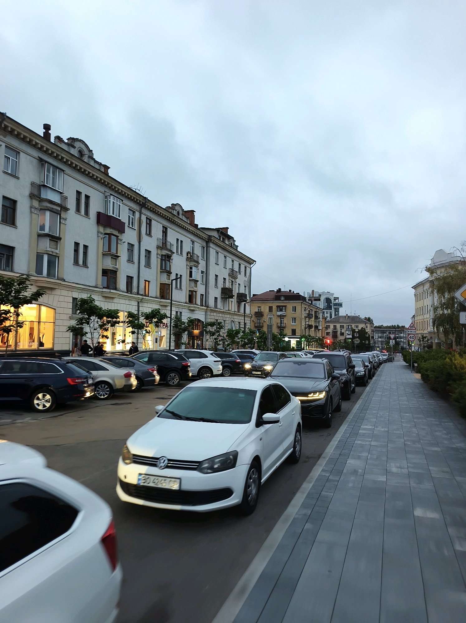The image shows a street scene in Ternopil, Ukraine, with a line of cars parked on the left and a sidewalk on the right. The buildings lining the street are multi-story, with architectural details suggesting an older European style. The sky is overcast, indicating a cloudy day, likely in the early morning or late afternoon given the artificial lighting from some storefronts.
Several cars are visible, including a white Volkswagen Polo in the foreground with a license plate that appears to be from Ukraine (BO 4245 CP). Other cars, mostly dark sedans and SUVs, are lined up behind it. Some of the parked cars are in designated parking spots along the curb, while others seem to be part of a traffic queue. There are trees planted along the sidewalk, and the ground is paved with cobblestones. A few people can be seen walking on the sidewalk and near the entrance of a building. One of the buildings has large illuminated windows, suggesting it might be a commercial establishment like a shop or cafe. A traffic sign indicating a speed limit of 40 km/h is visible on the right side of the street.