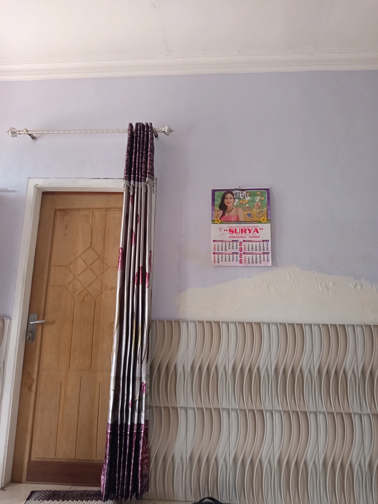 The image shows an indoor scene with a door and a wall decorated with a calendar and patterned panels. A light purple wall is visible at the top, with a white ceiling molding. A curtain rod with decorative ends is mounted on the wall, with a dark purple patterned curtain hanging from it. To the right of the curtain, a calendar is attached to the wall. The calendar features a woman's image and promotional text, and a portion of the calendar grid is visible, showing dates. Below the wall with the calendar, there is a section of decorative panels with a wavy, textured pattern in shades of cream and light brown. A wooden door with a metal handle is visible on the left side of the image. The door has a diamond-shaped pattern carved into its surface. A small rug is placed on the floor in front of the door. The overall impression is of a residential interior, possibly in the city of Rowo Indah, Indonesia, given the context.