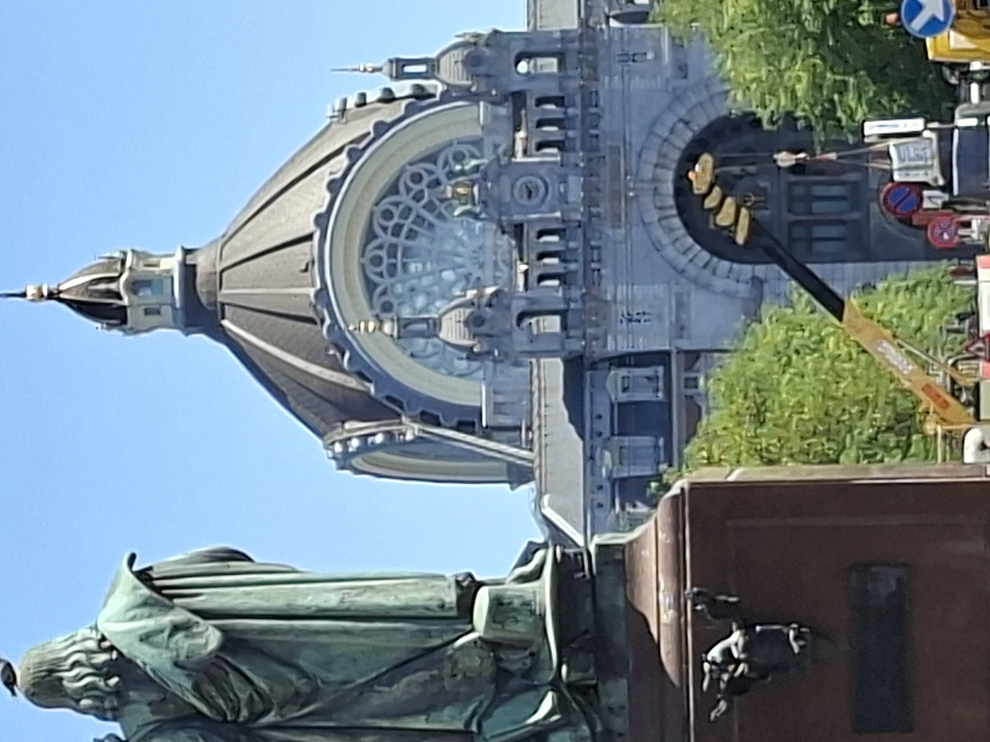 This is a photograph taken from street level, looking towards a large, ornate building with a prominent dome. In the foreground on the left, a portion of a large bronze statue is visible, showing the back of a robed figure with a bird perched on its head. The statue appears to be on a reddish-brown pedestal, with some decorative elements attached to the side of the pedestal.
Behind the statue and to the right, the ornate facade of the building dominates the background. This building features classical architectural elements, including arched windows, decorative stonework, and smaller domed turrets flanking the main dome. The main dome is dark gray and segmented, with a pointed finial at its apex. A large, intricate circular window is visible within the building's facade below the dome.
The scene is set outdoors under a clear blue sky, suggesting a sunny day. Lush green trees are visible in the mid-ground, partially obscuring the lower sections of the building. In the lower right quadrant of the image, a yellow construction crane is partially visible, with a hook and some suspended objects. Traffic signs, including a red circular sign with a blue inner circle and a white diagonal bar, and a blue circular sign with a white arrow pointing left, are also present on the street.
The building in the background is the Antwerp Central Station, also known as Middenstatie, a historically significant railway station in Antwerp, Belgium. The statue in the foreground is likely a monument located in the vicinity of the station.