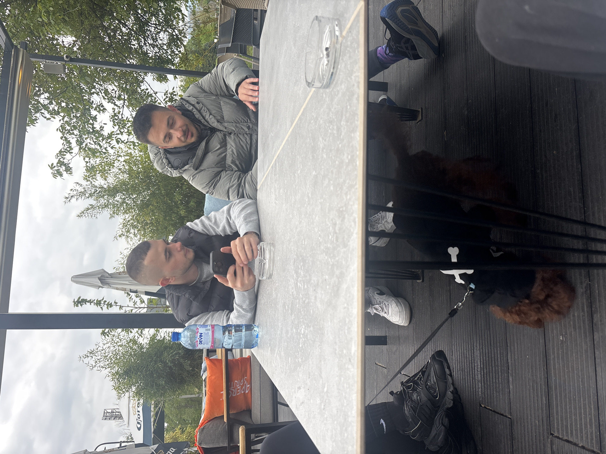 Two men and a small, fluffy brown dog are seated at an outdoor cafe in Plovdiv, Bulgaria, on what appears to be an overcast day. The setting features dark wooden decking and black metal railings.
One man, wearing a grey puffer jacket, rests his head on his arms, which are folded on a light grey, concrete-like table. A glass ashtray containing a cigarette butt is also on the table near him. The second man, dressed in a black vest over a grey long-sleeve shirt, is actively looking at a black smartphone he holds with both hands. A plastic bottle of "DEVÍN" branded water is placed near him on the table. Behind him, an orange cushion with partial text, likely "APEROL SPRITZ," is visible on a chair.
Under the table, the small brown dog is tethered by a leash, lying calmly on the decking. Various shoes, including black running shoes, white sneakers, and blue and purple sneakers, are scattered beneath the table, suggesting multiple individuals or items.
The background shows lush green trees against a cloudy sky. Further in the distance, a large structure and white umbrellas, one bearing the "CORONA" logo, indicate a commercial or recreational area. An outdoor heater or security camera is mounted above the seating area. The overall atmosphere is casual and relaxed.