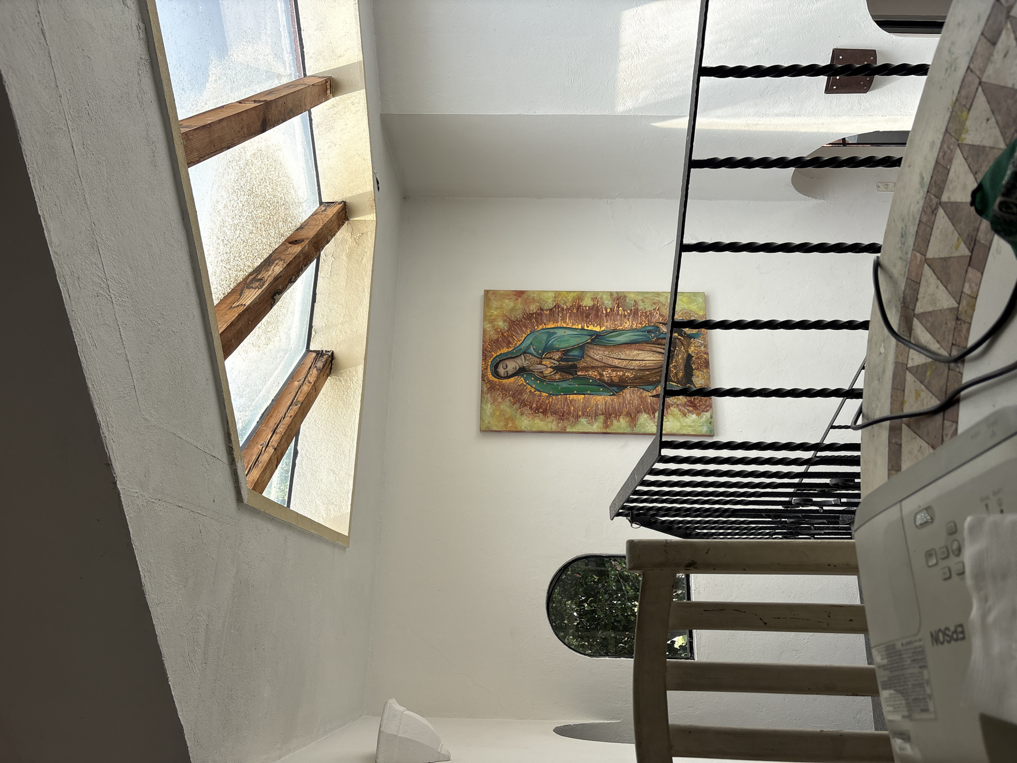 The image shows an interior view of a building in Puebla, Mexico, characterized by white walls and a unique architectural style. A prominent feature is a window with wooden beams, allowing natural light to enter the space. On the wall, there is a painting of the Virgin of Guadalupe. A metal staircase is visible, along with a wooden chair and a white Epson printer in the foreground. A circular window is present on the wall, providing a glimpse of greenery outside.