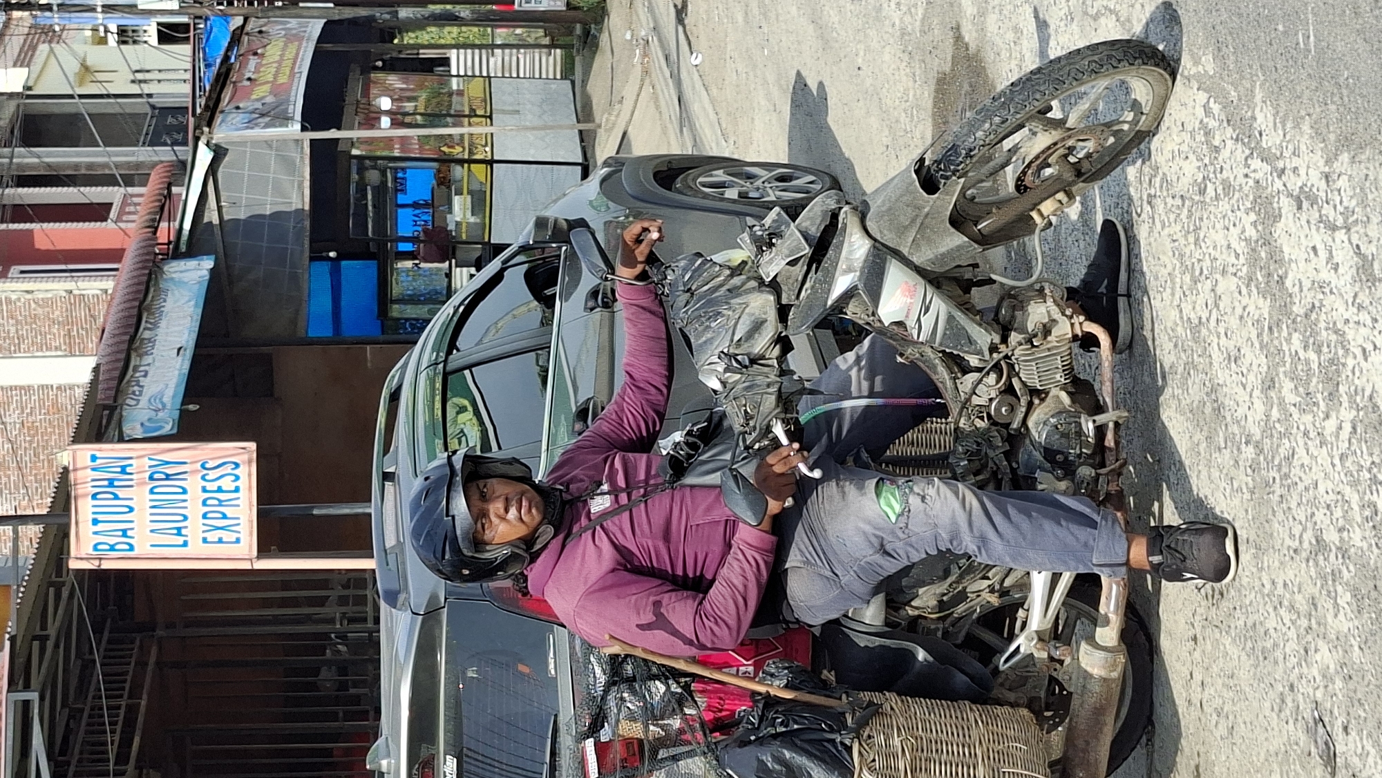 A man wearing a maroon hooded sweatshirt, dark pants, and a black helmet is seated on a dirt-covered motorcycle. He is looking towards the camera. The motorcycle is heavily loaded with bags and has a wicker basket attached to the rear. Behind the motorcycle, a dark gray SUV is parked. In the background, a sign with blue lettering that reads "BATUPHAT LAUNDRY EXPRESS" is visible, along with buildings and street activity. The scene appears to be on a street in Lhokseumawe, Indonesia.