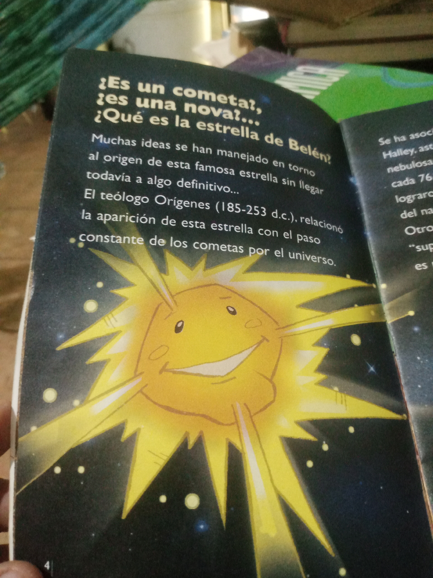 The image shows an open book with a prominent illustration of a smiling yellow sun-like character with sharp rays extending outwards against a dark, starry background. The text on the left page is in Spanish and discusses comets, stars, and the star of Bethlehem. The text on the right page is partially visible, also in Spanish, mentioning "Se ha asocia" and "Halley, ast". A portion of a green object is visible at the top right of the frame, likely part of another book or item. The lighting suggests it is indoors, with the book lying on a surface. The image does not contain any information that would allow for the identification of its location in Santa Ana, Venezuela.