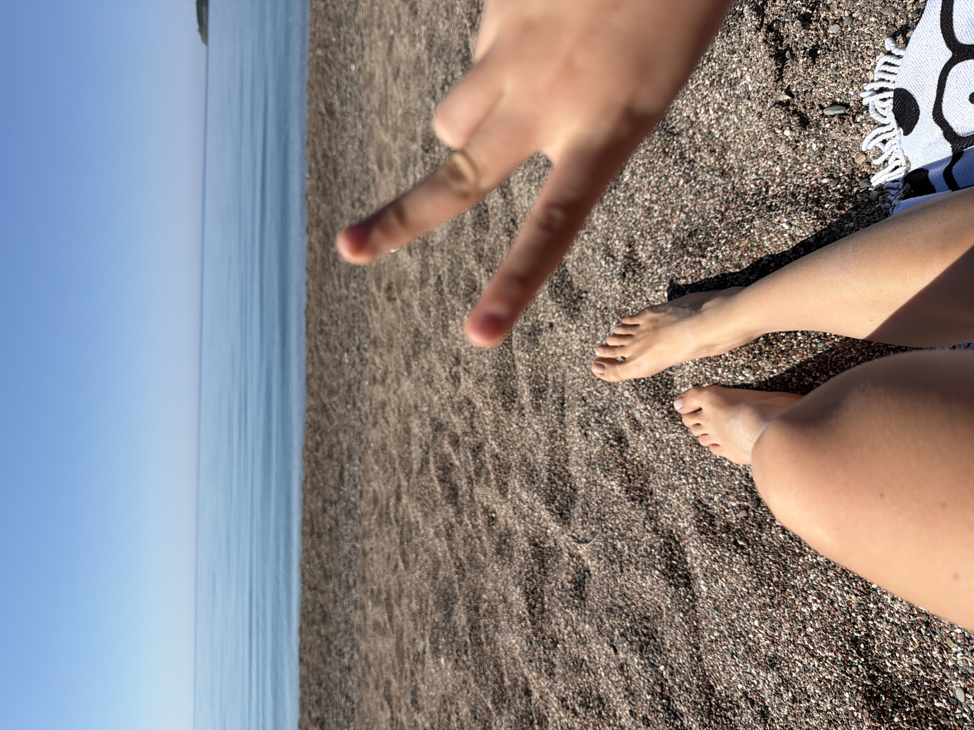 The image, taken from a first-person perspective, captures a serene beach scene in Rafailovići, Montenegro. The primary subjects are the legs and feet of a person, likely the photographer, resting on a pebble beach. In the foreground, a hand makes a peace sign towards the camera, conveying a relaxed and happy mood.
The setting is a sunny day on a dark pebble beach, typical of the Adriatic coast. Bright sunlight casts distinct shadows from the legs onto the small, dark stones, suggesting it is late morning or early afternoon. Part of a white towel with a black, possibly abstract or tribal, pattern is visible on the right side of the frame, tucked under a leg.
Beyond the beach, the calm, light blue waters of the Adriatic Sea extend to a distant horizon, meeting a clear, vibrant blue sky. Faint details of a faraway coastline or landmass can be seen where the sea meets the sky. The overall impression is one of leisure and enjoyment during a peaceful beach holiday. There is no visible text in the image.