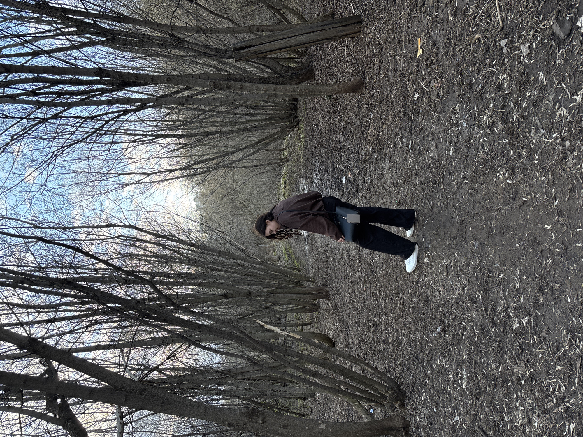 The image captures a young person standing on a dirt path lined with numerous bare deciduous trees, suggesting a setting in late autumn, winter, or early spring in Perm, Russia. The individual, who appears to be a woman with long, dark, wavy hair, is dressed in a brown jacket, black pants, and white sneakers, carrying a dark crossbody bag. She is positioned towards the right side of the path, looking downwards or away from the camera.
The path itself is unpaved, covered in dark soil, scattered dry leaves, and small twigs, with visible damp or muddy patches. Tall, slender trees with intricate networks of bare branches line both sides of the path, forming a natural tunnel or avenue. Through the overhead branches, a bright, partly cloudy sky is visible, indicating daytime. In the middle distance to the right, a weathered wooden structure, possibly a discarded plank or fallen log, rests on the ground amidst the trees. The overall scene conveys a quiet, somewhat somber atmosphere typical of the colder seasons in a natural, wooded environment. No visible text is present in the image.
